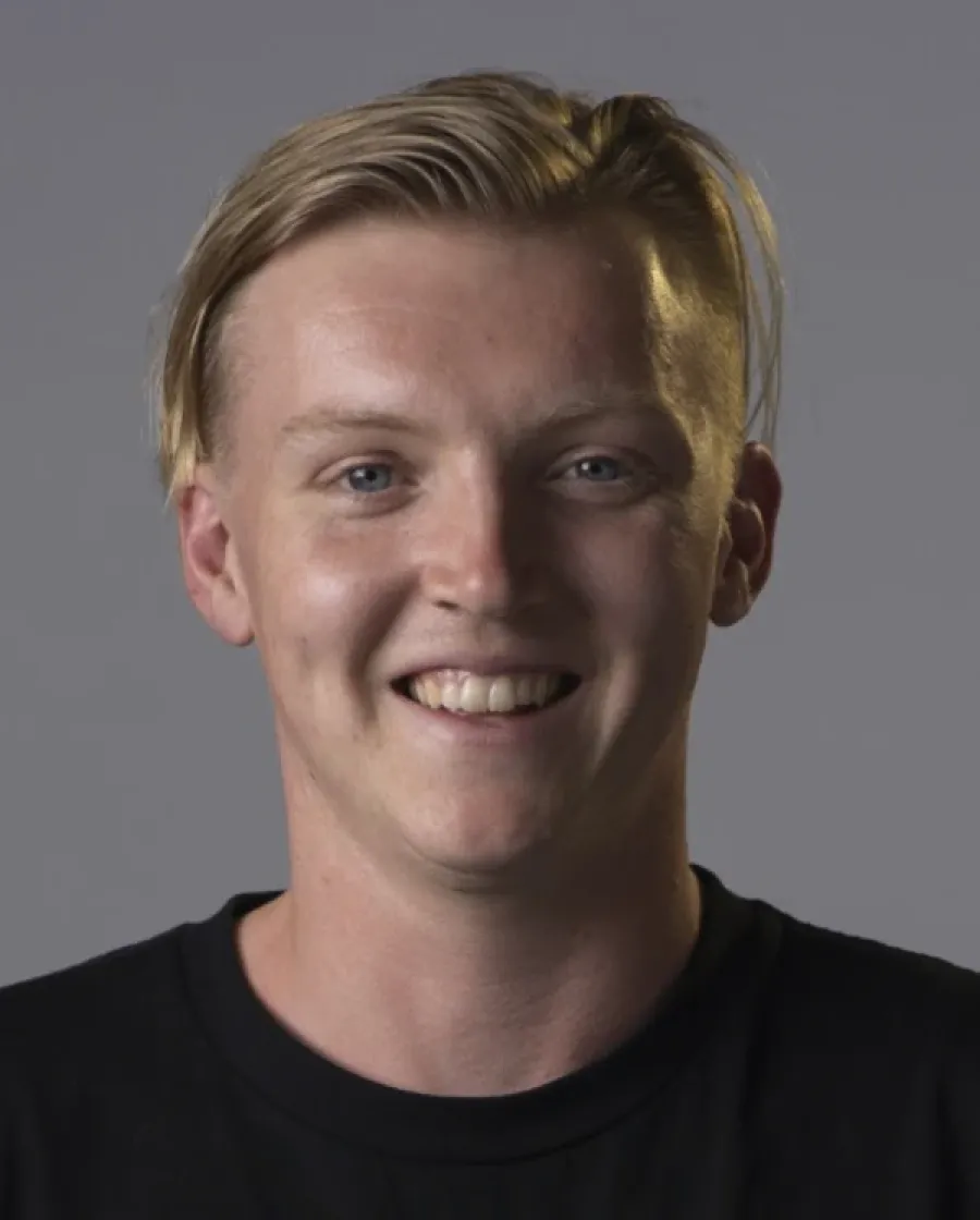 Close-up portrait of Max who is a smiling man with blonde hair swept to the side, wearing a black shirt, against a plain gray background.