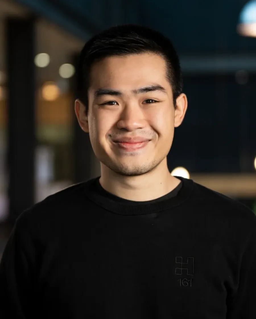 Portrait of Cedric who has short dark hair and is smiling slightly, wearing a black shirt with a subtle Halter logo, photographed indoors with blurred lights in the background.