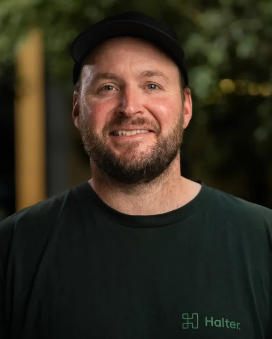 Portrait of Mark who is a bearded man wearing a black cap and a green Halter-branded shirt, smiling at the camera in a warmly lit indoor space with greenery in the background.