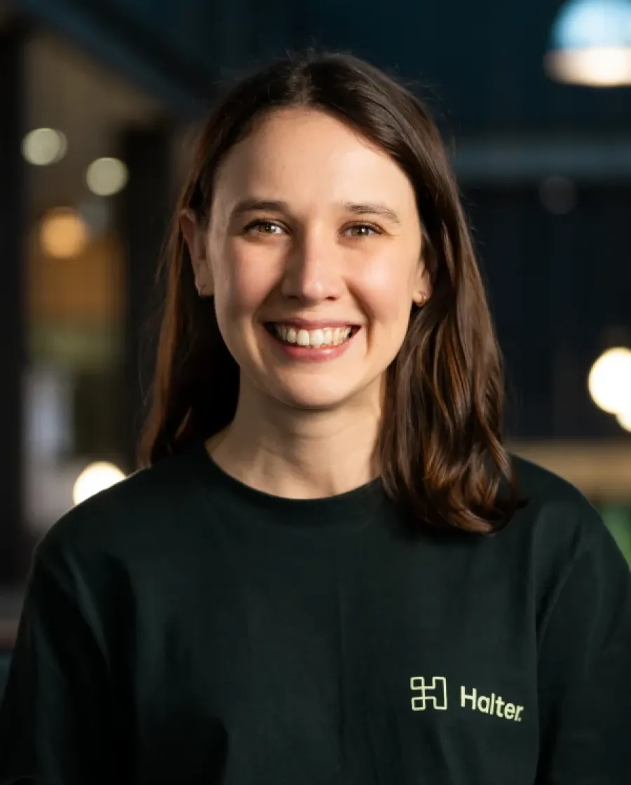 Portrait of Dani who is smiling woman with medium-length dark brown hair, wearing a black Halter-branded shirt, posing in a warmly lit indoor environment.
