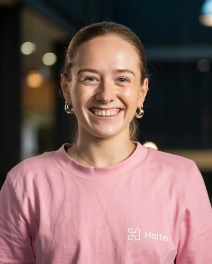 Portrait of V Gregan. She is a woman with her hair tied back, smiling brightly, wearing a pink Halter-branded shirt, photographed indoors with soft lighting.