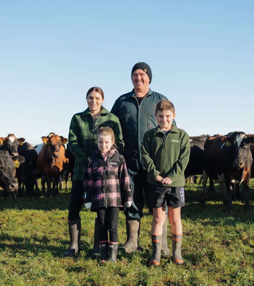 A farming family of four stands together in a grassy pasture, smiling at the camera with a herd of cows behind them on a clear, sunny day.