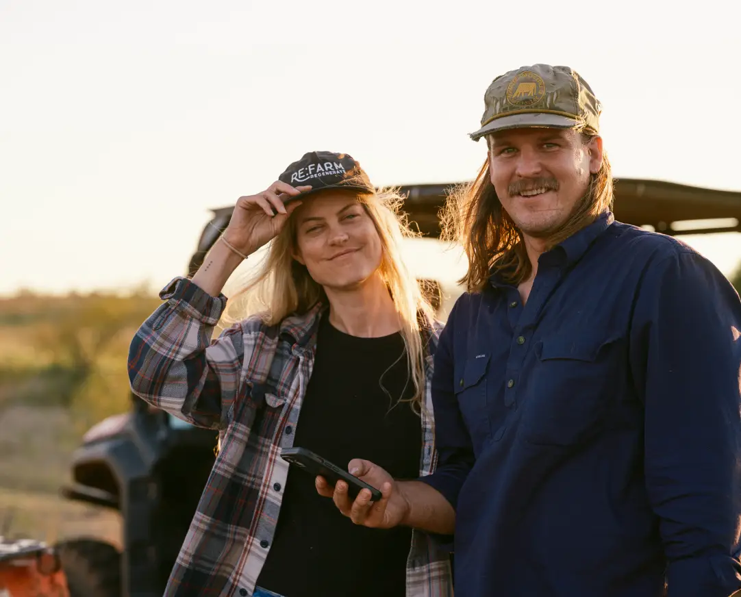 Two smiling Halter ranchers stand outdoors at sunset, one holding a phone and the other tipping their hat, with a utility vehicle in the background.