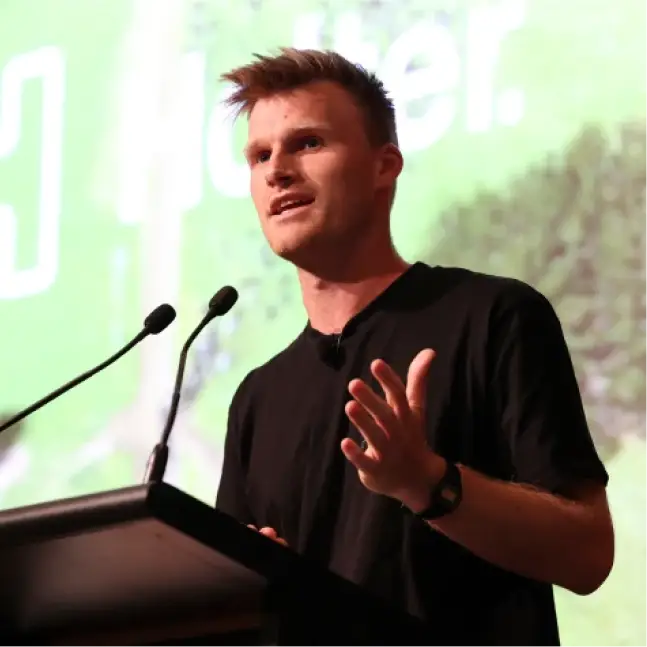 Craig Piggott speaks on stage at a podium with two microphones, gesturing with one hand