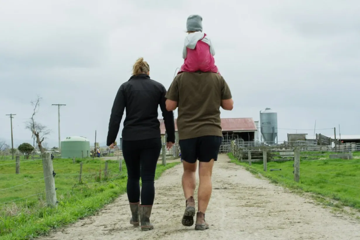 A woman and man walk down a gravel farm track, viewed from behind. The man carries a young child on his shoulders, and farm buildings and fencing are visible in the background.