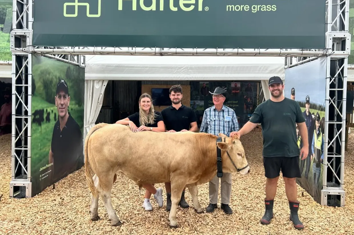 Some Halter team members & a farmer stand behind a light brown cow wearing a Halter Collar at a Field Days booth for Halter. 