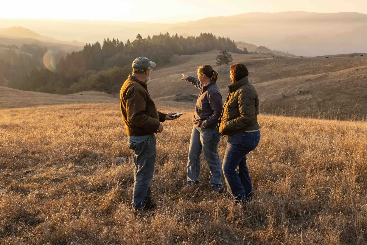 Three ranchers stand in a dry, grassy landscape at sunset, with one woman pointing toward the hills and trees in the distance.
