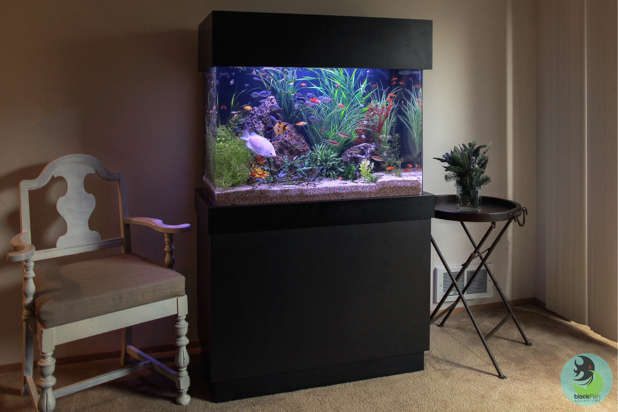Large aquarium with colorful fish and plants on black stand beside a wooden chair and round table with flowers.