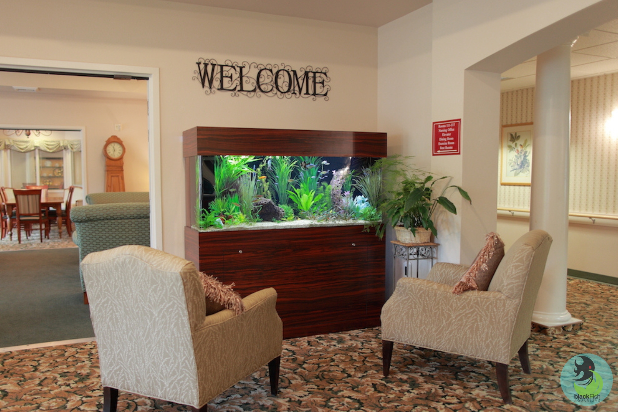 Two beige armchairs face a large wood-paneled aquarium in a cozy room with patterned carpet.
