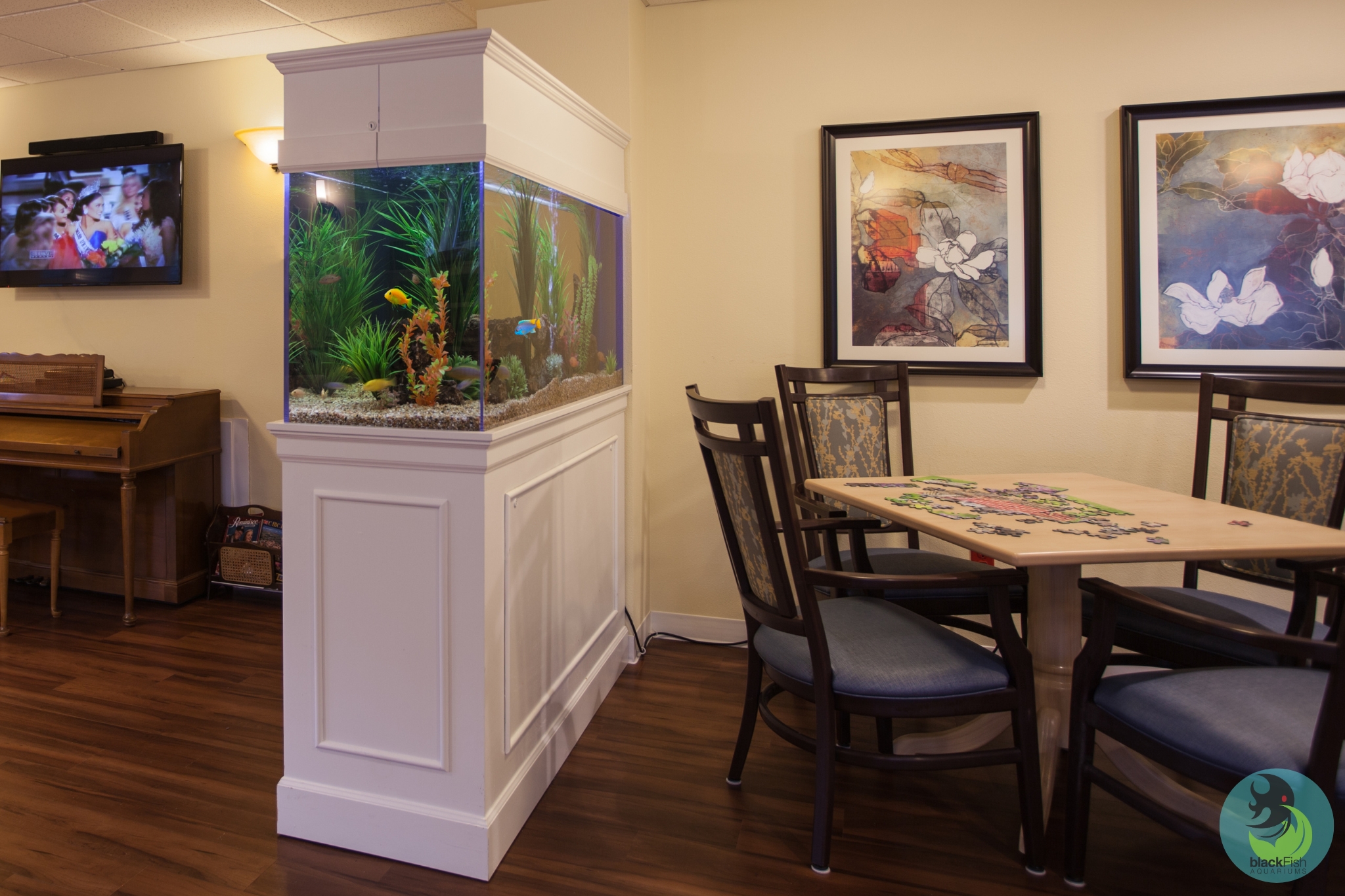 Indoor room with a large white aquarium, wooden piano, a table with puzzle pieces, and floral wall art.