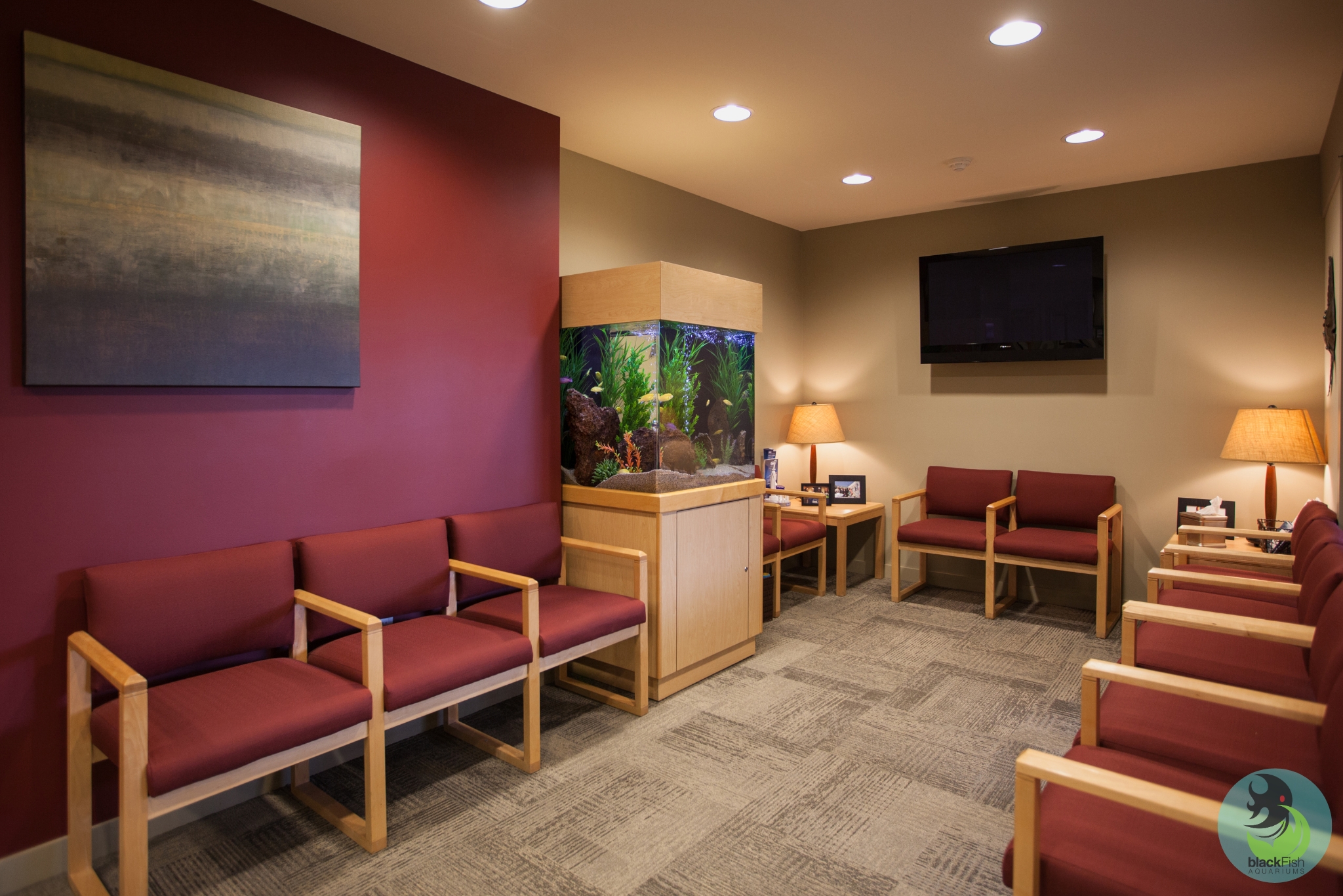 Waiting room with maroon chairs, aquarium, lamps, a TV on beige walls, and gray carpeted floor.