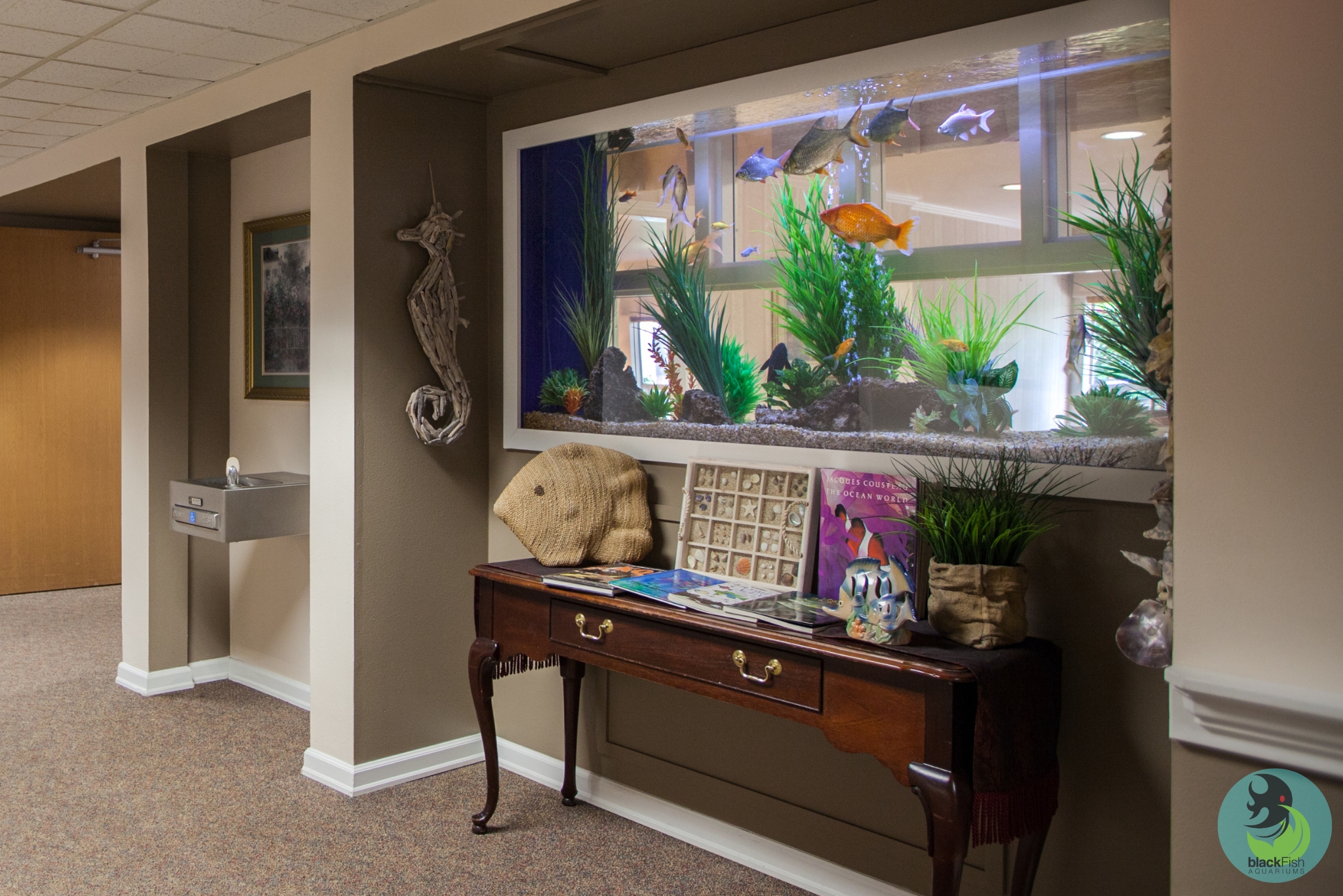 Large wall-mounted fish tank with colorful fish above a wooden table with ocean decor and books.