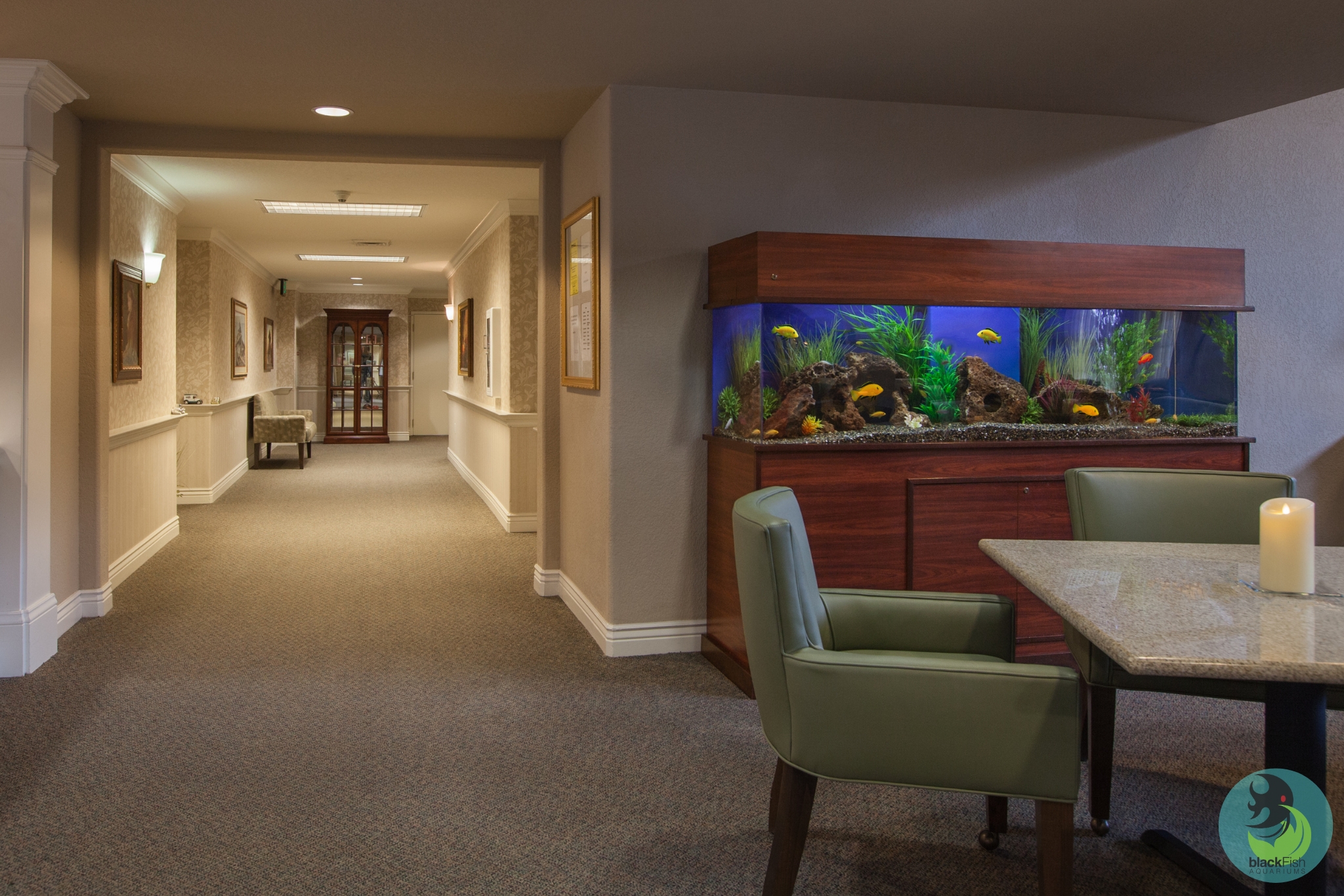 Hallway with carpet and paintings, featuring a large fish tank with yellow fish near a table and chairs.
