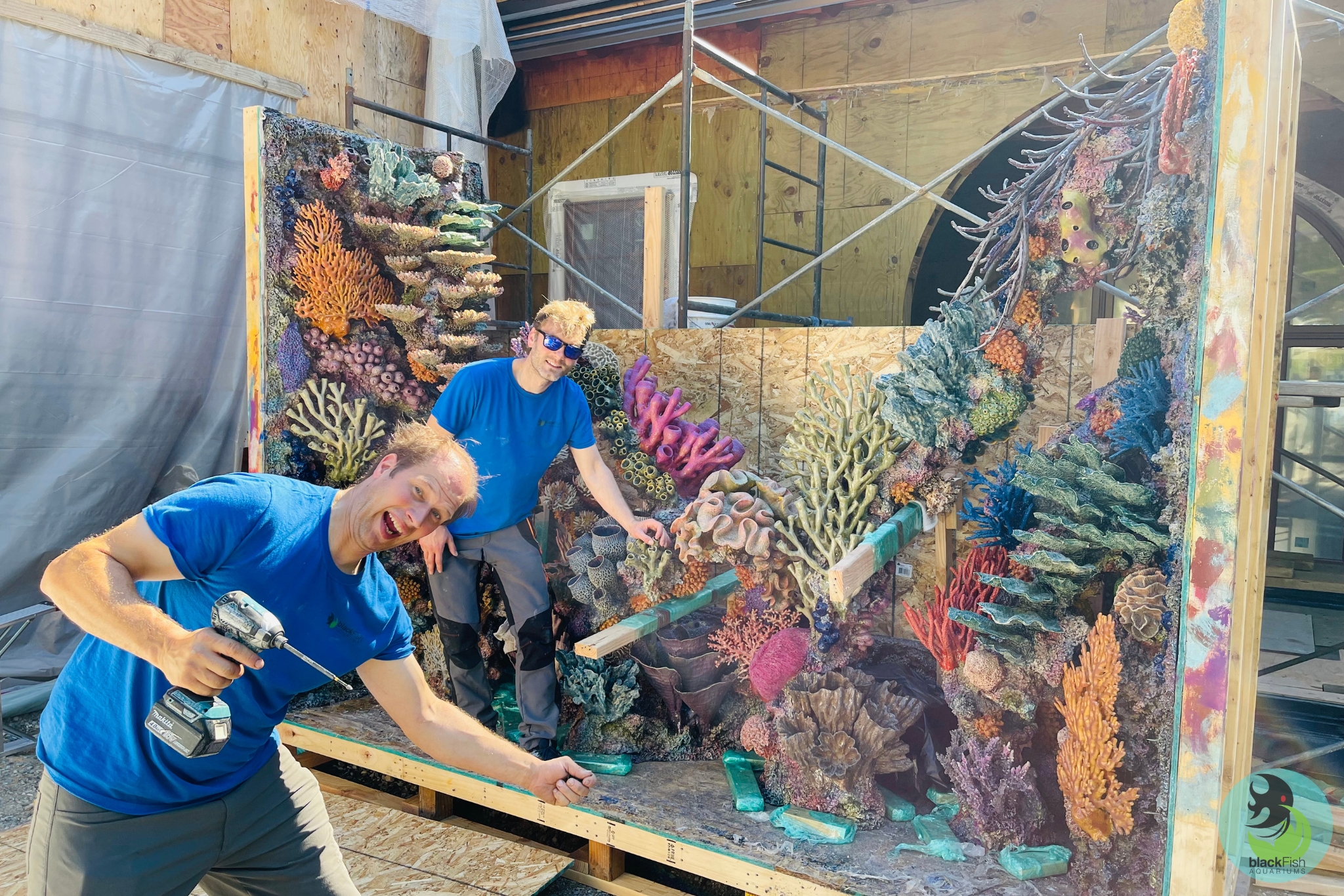 Two men in blue shirts assembling a large colorful artificial coral reef display inside a wooden frame.