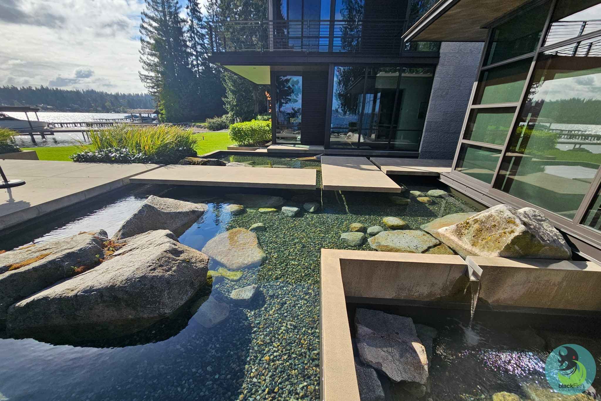 Modern house with a clear water pond featuring large rocks and concrete stepping stones near a lake.