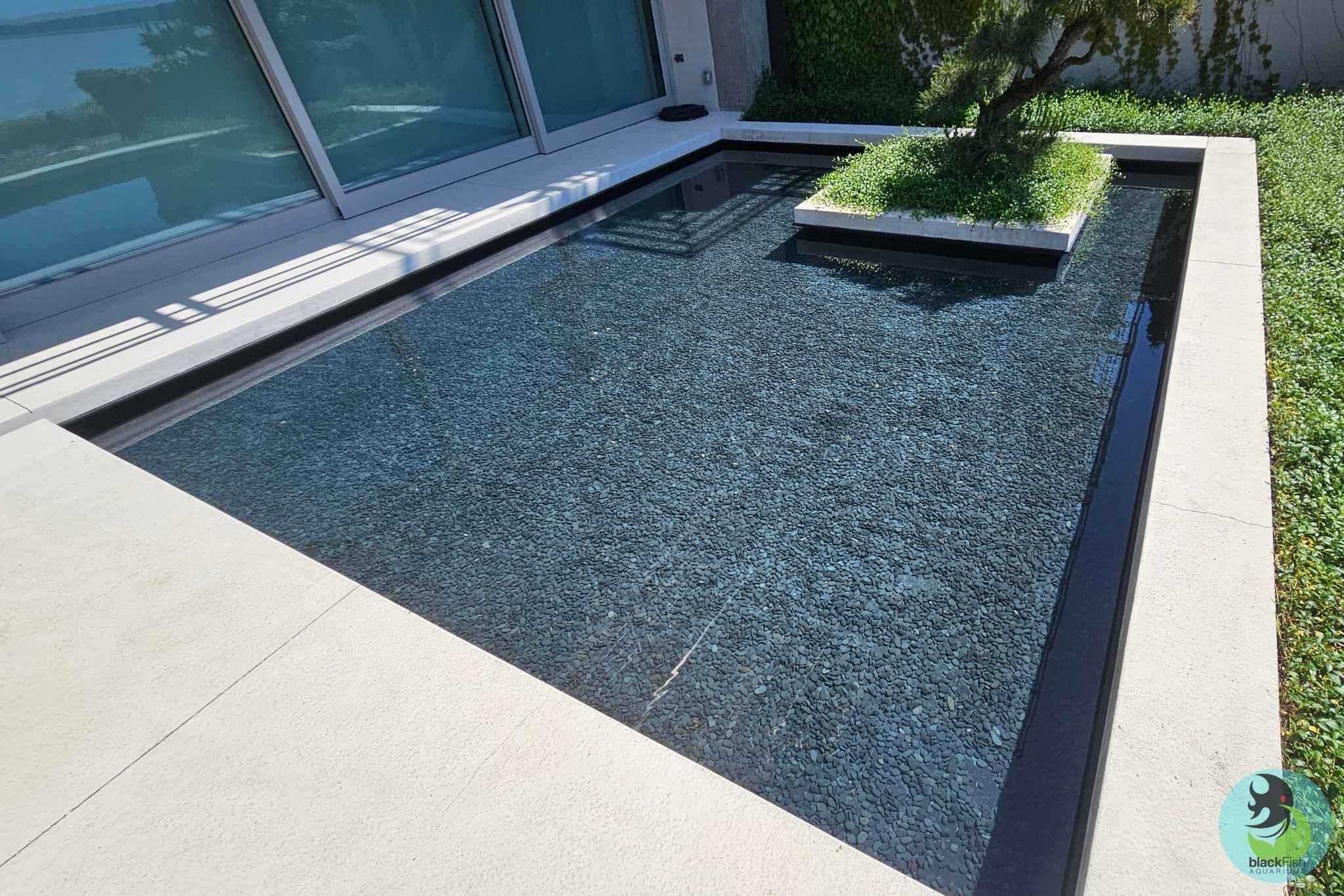 Modern outdoor water feature with clear water, black stones, and a small tree on a raised platform.