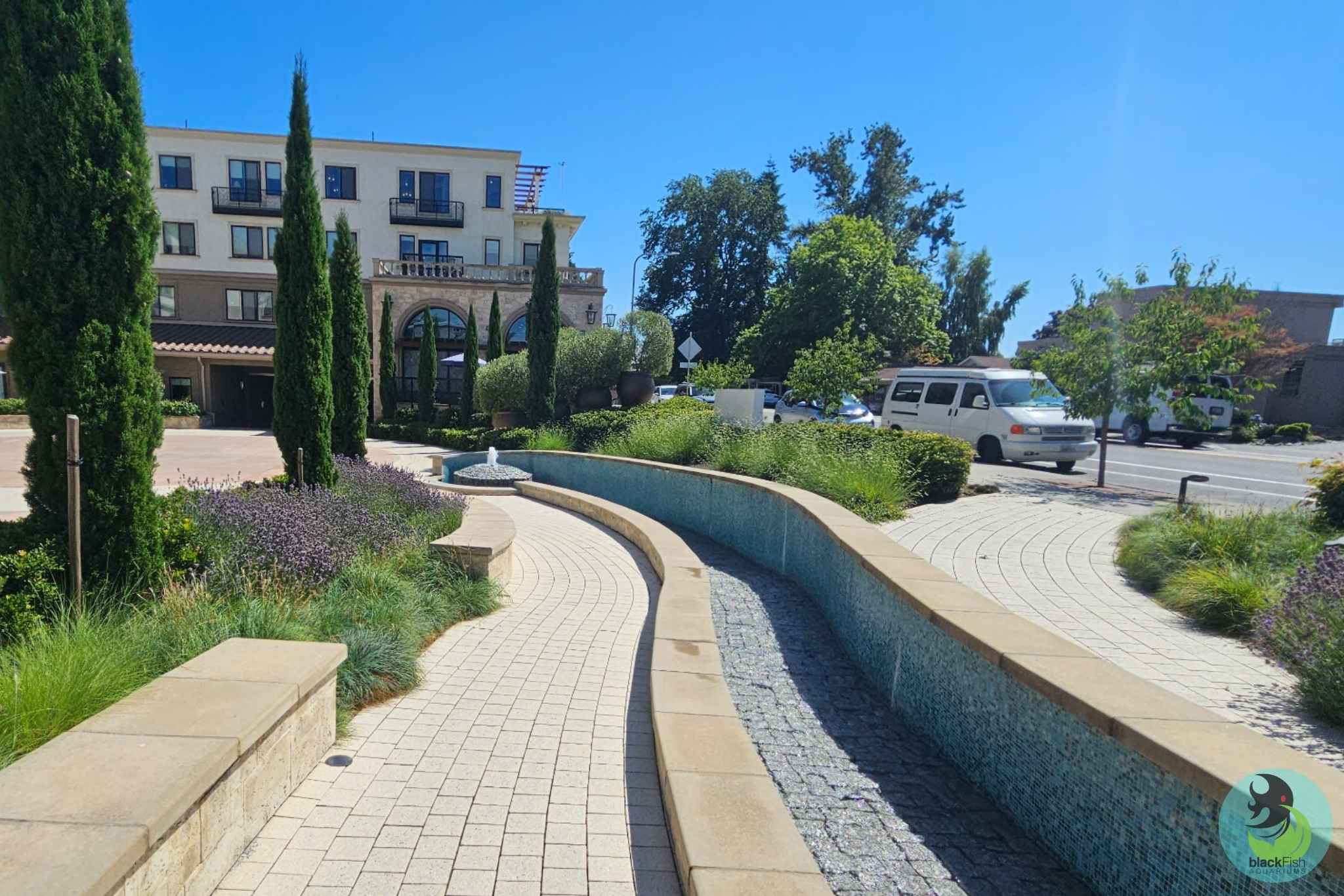 Curved tiled walkway with a dry stone-lined water channel and benches beside greenery and trees.