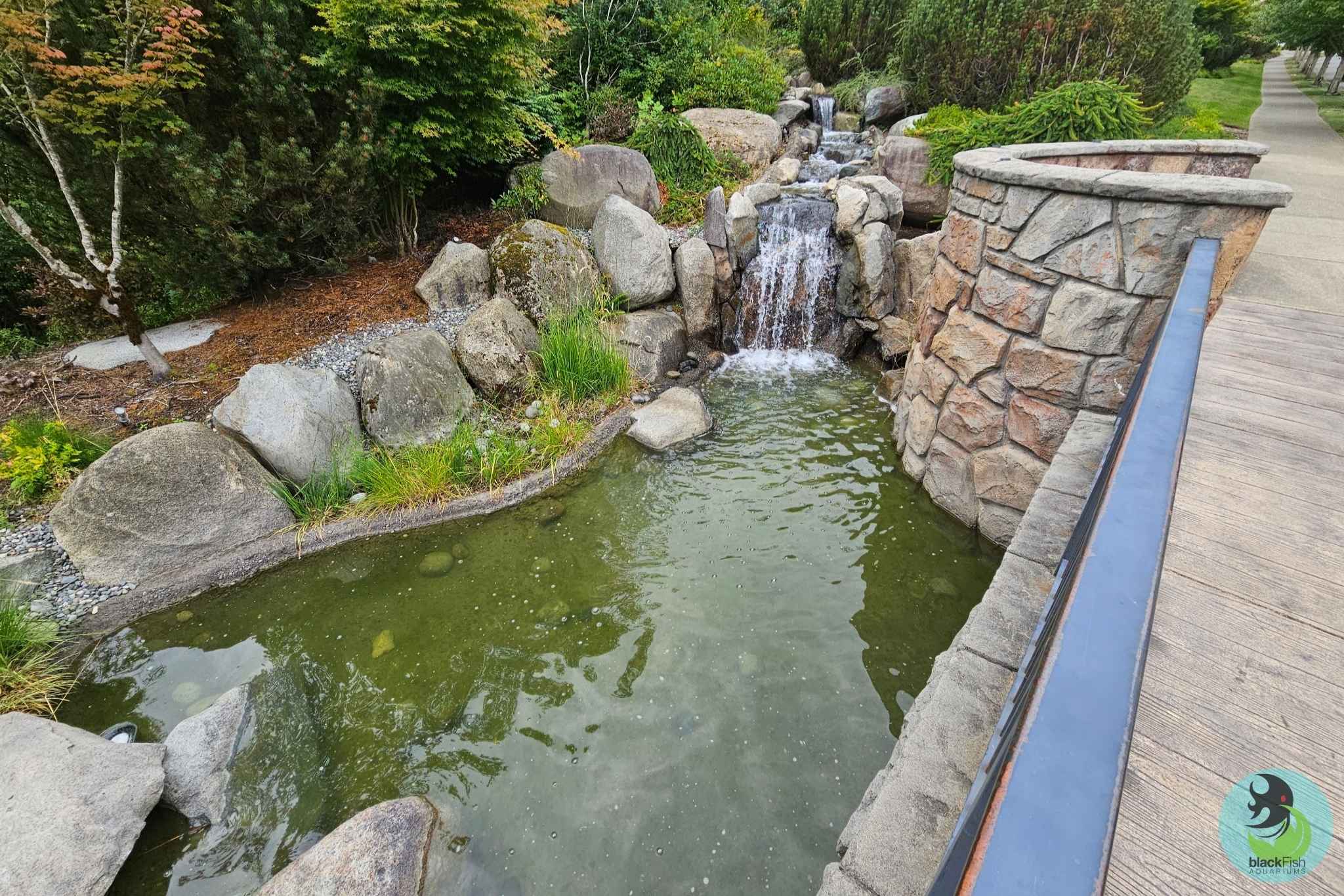 Small waterfall flowing into a pond beside a stone wall and wooden walkway surrounded by greenery.