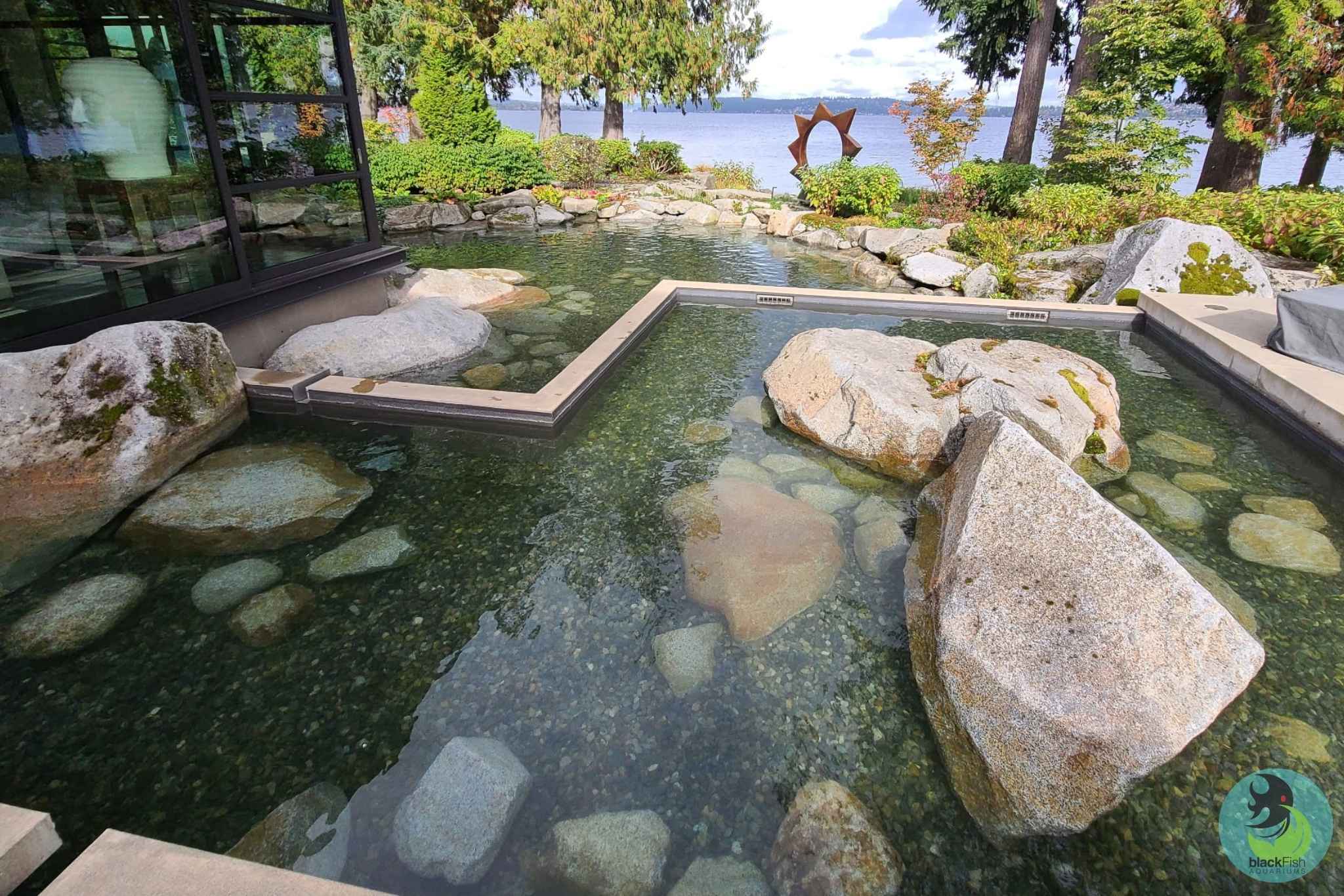 Clear outdoor pond with large rocks bordered by landscaping and lake view beyond trees.