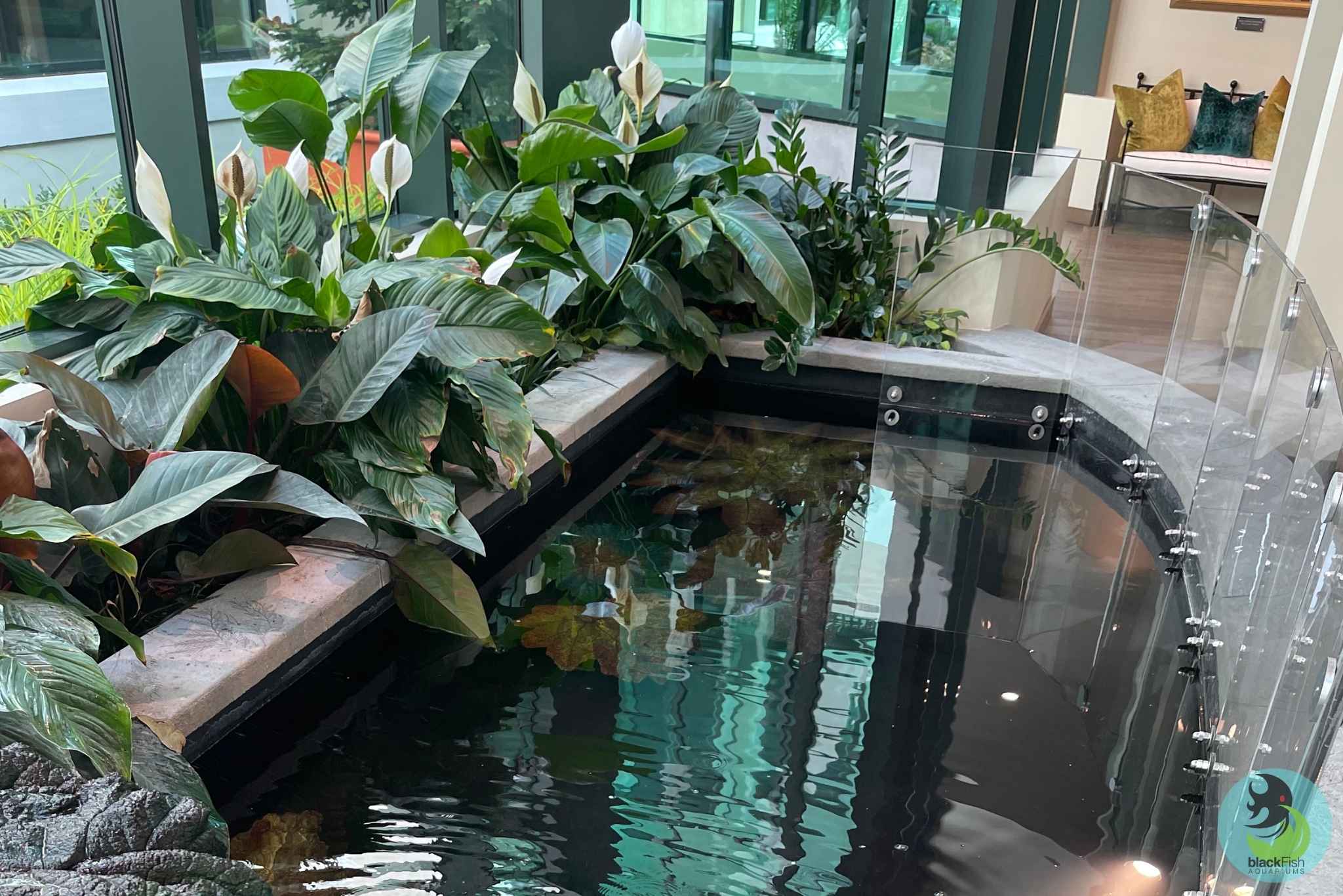Indoor pond with clear water bordered by leafy green plants and a glass safety barrier.