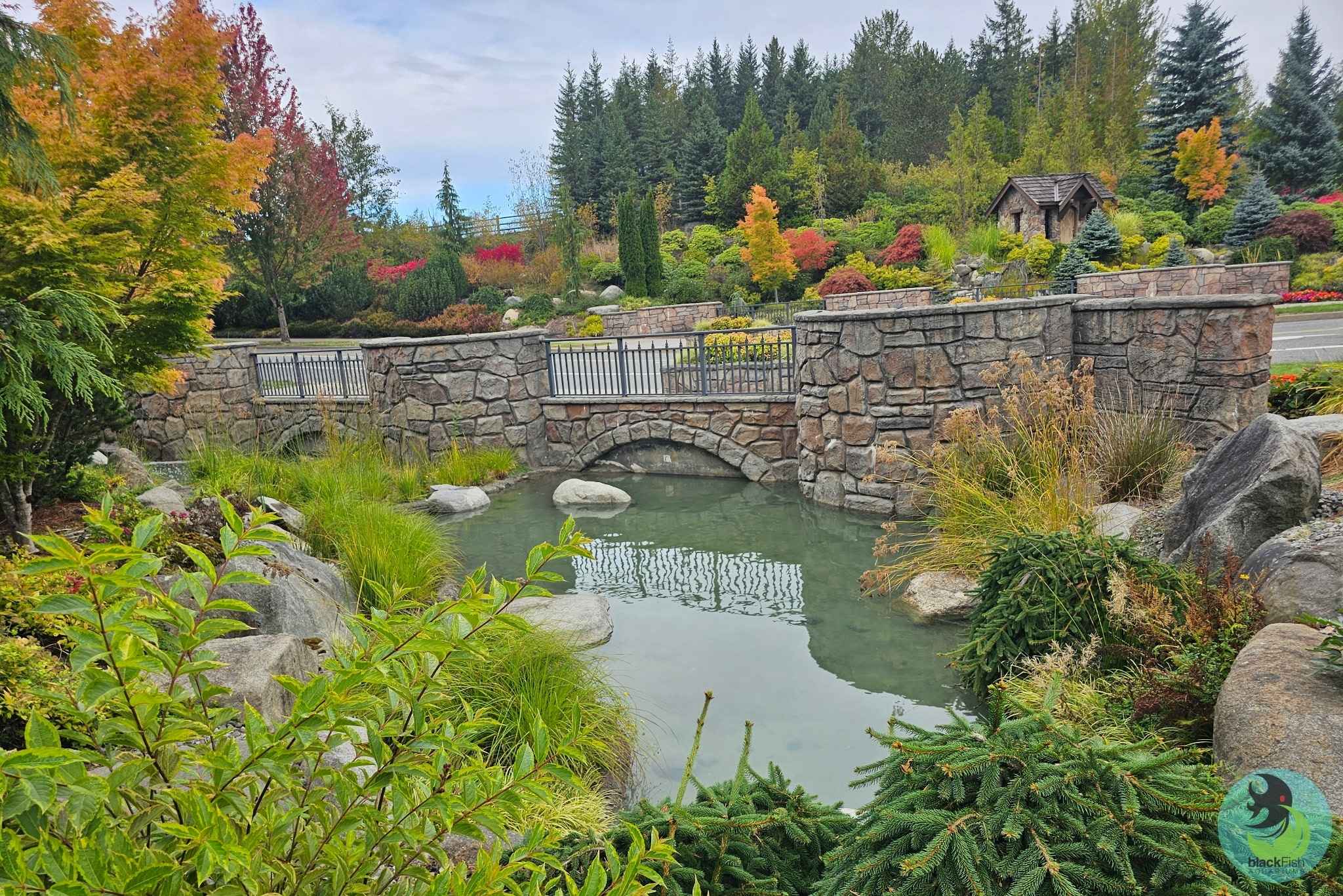 A stone bridge over a clear decorative pond surrounded by autumn trees and varied greenery.