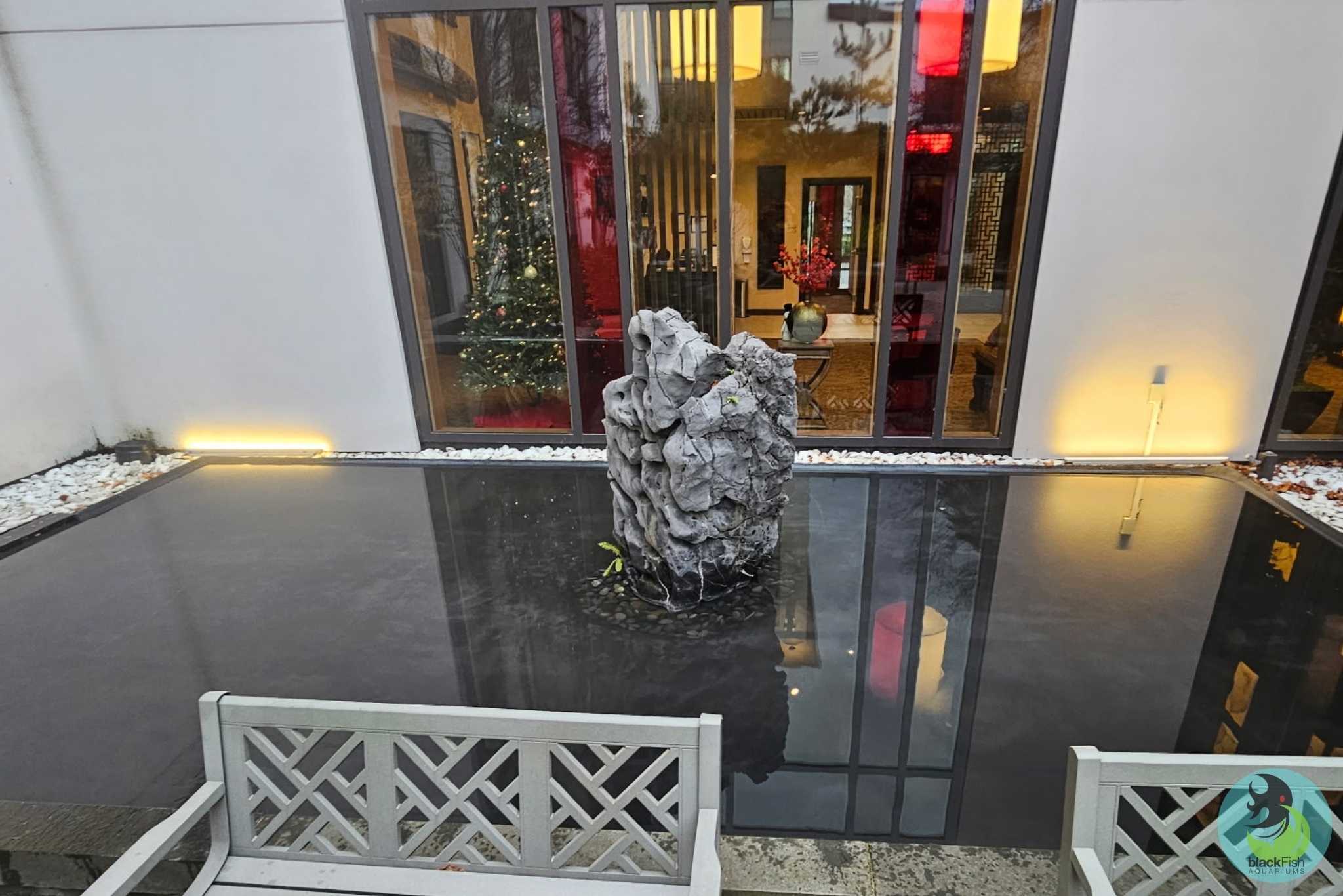 Modern courtyard pond with rock sculpture center, white benches, and indoor Christmas tree reflected.