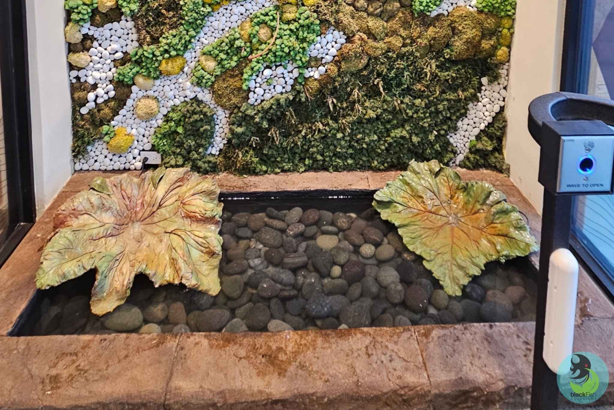 Indoor water feature with lotus leaf sculptures, clear water over stones, and a green moss wall behind it.