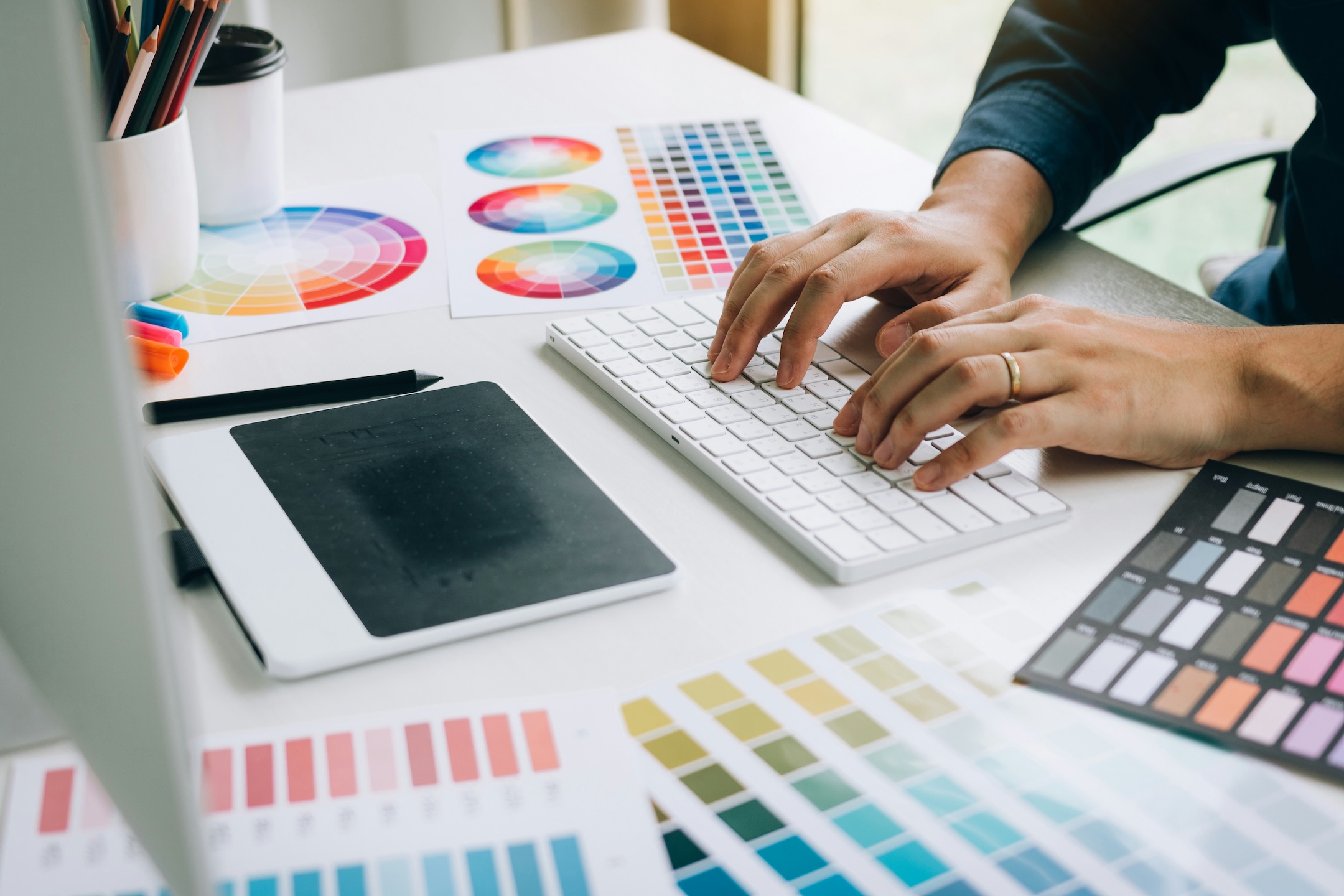 Hands typing on a keyboard surrounded by color swatches, color wheel charts, and a drawing tablet.