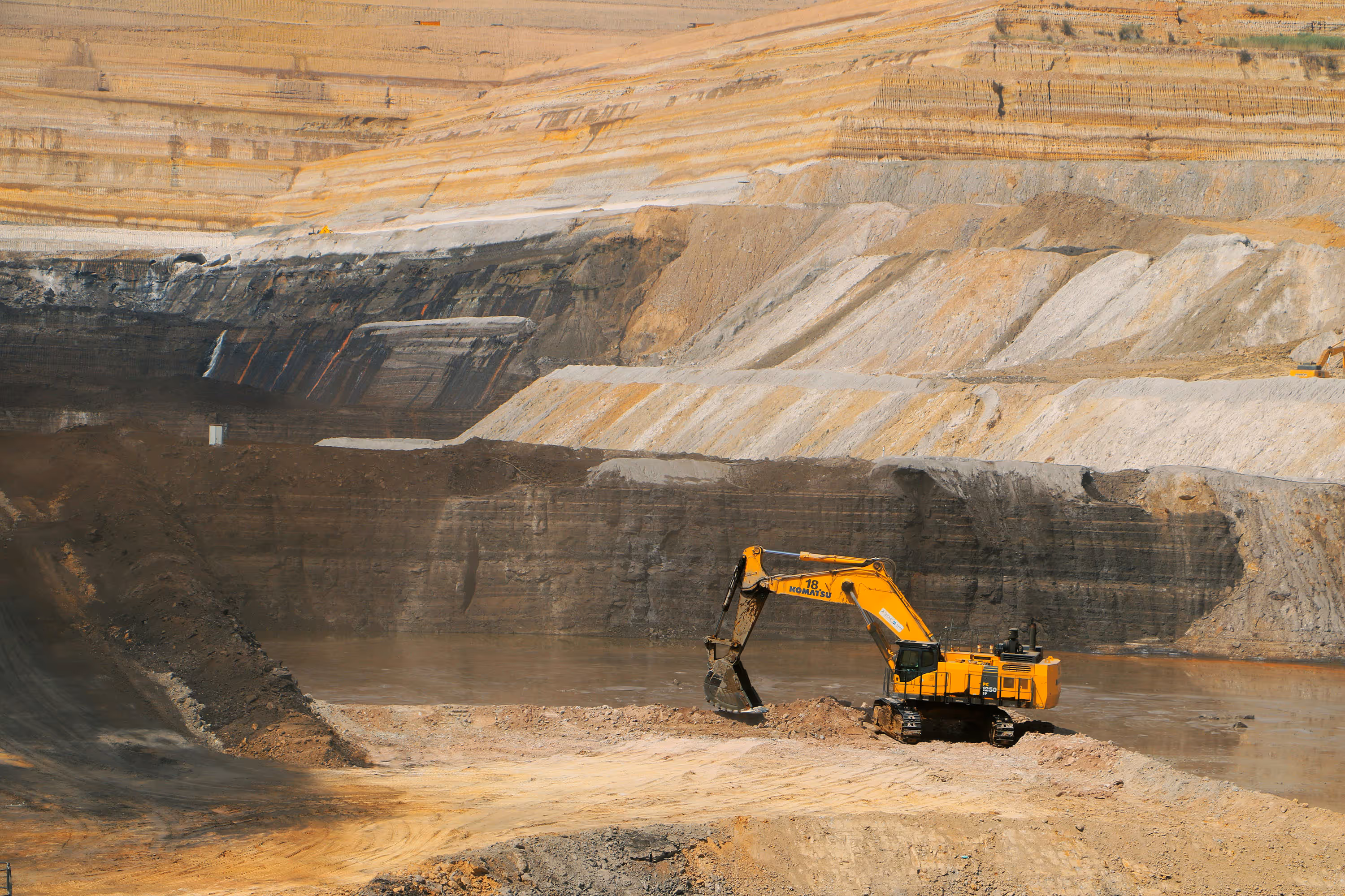 Large yellow excavator working in a deep open-pit mine with layered earth and rock walls.