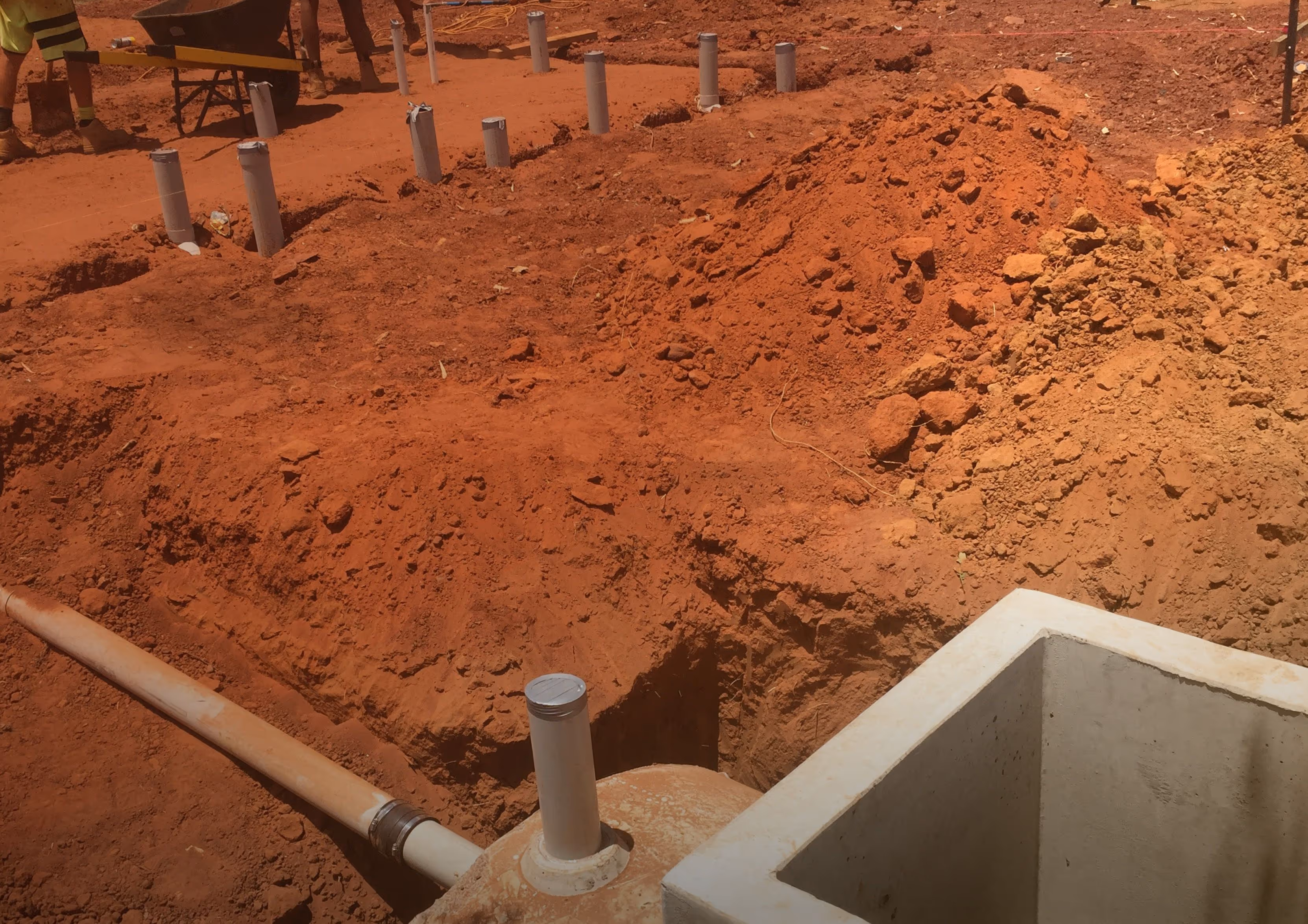 Construction site with exposed red soil, PVC pipes installed in trenches, and a concrete utility box.