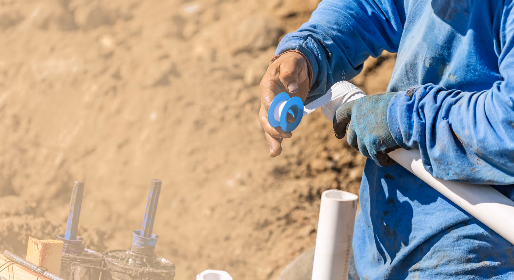 Worker wearing blue gloves and shirt wrapping white pipe thread tape around a PVC pipe at a construction site.