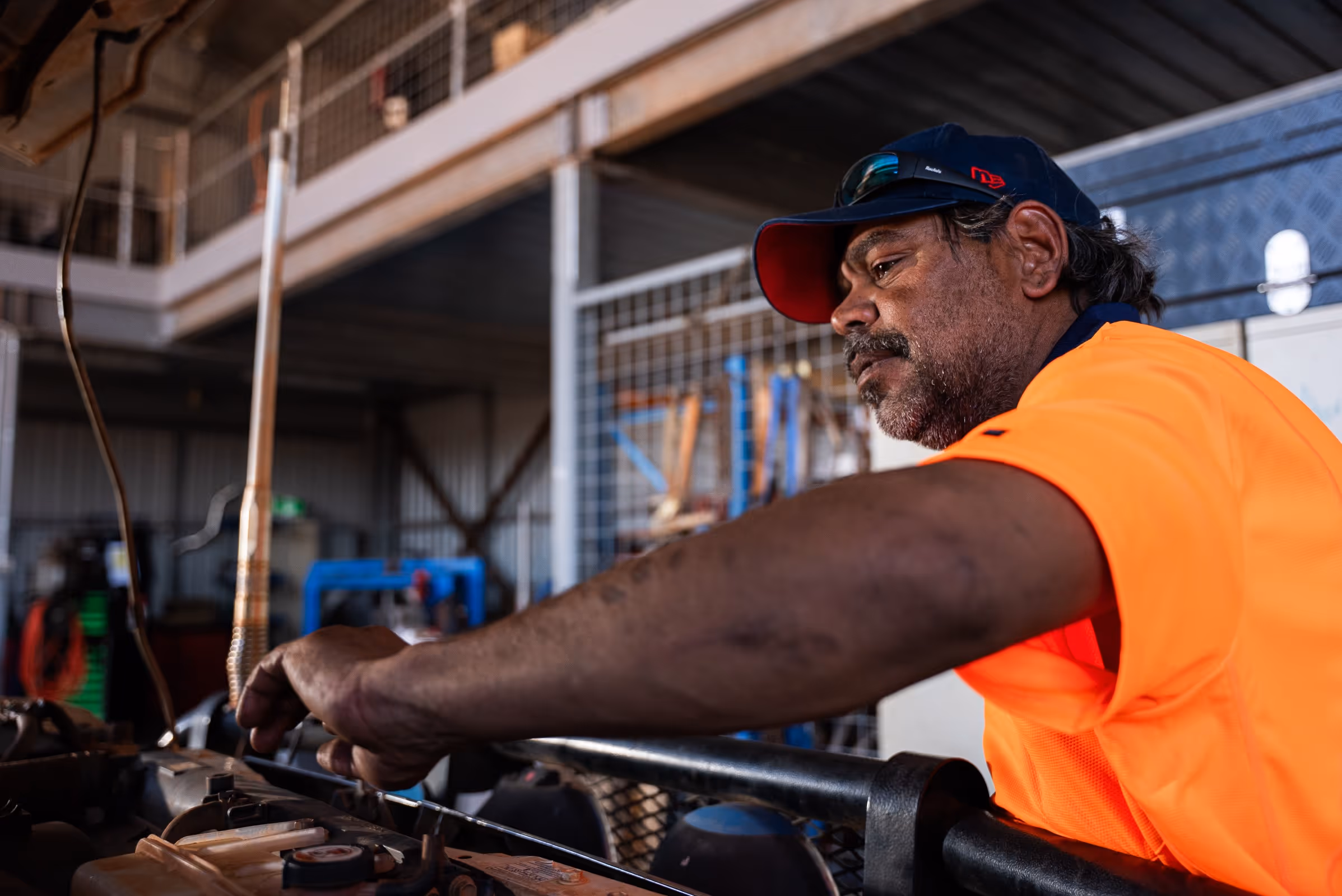 Man in an orange high-visibility shirt and cap working on a mechanical part in an industrial workshop.