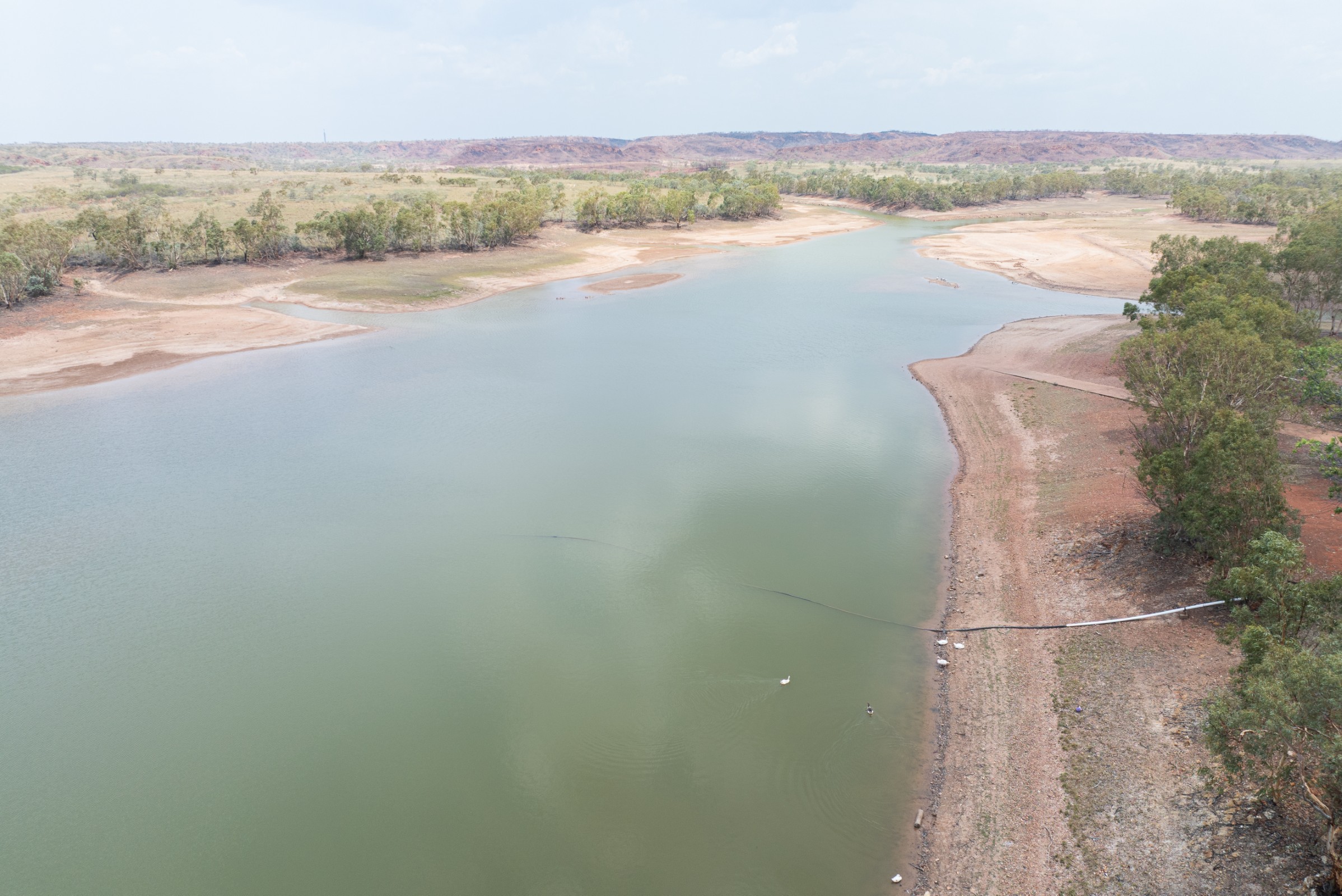 A wide river with calm green water bordered by dry, sparsely vegetated banks and scattered trees under a cloudy sky.