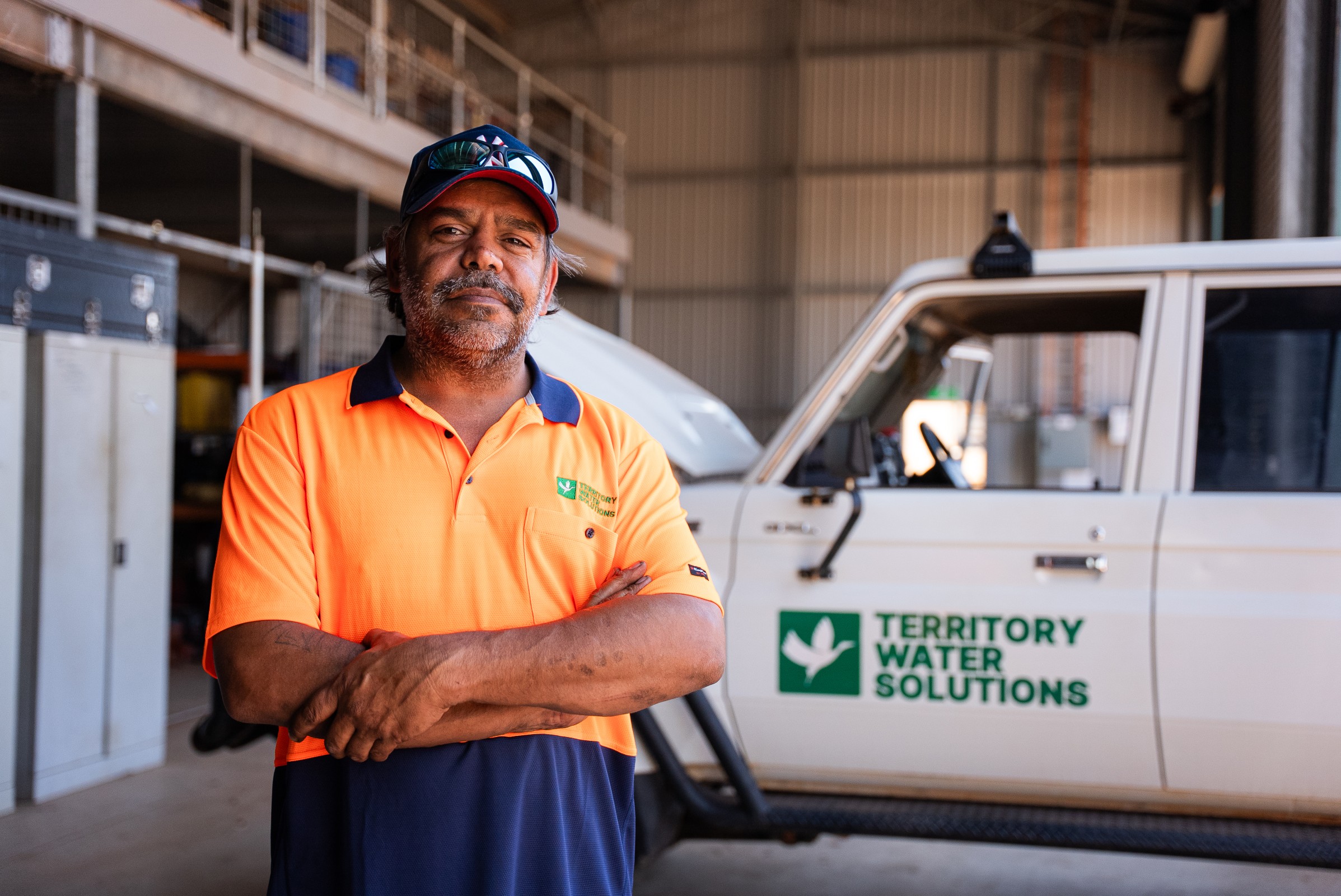 Man in an orange and navy Territory Water Solutions uniform standing with arms crossed inside a warehouse, next to a white vehicle with the company logo.