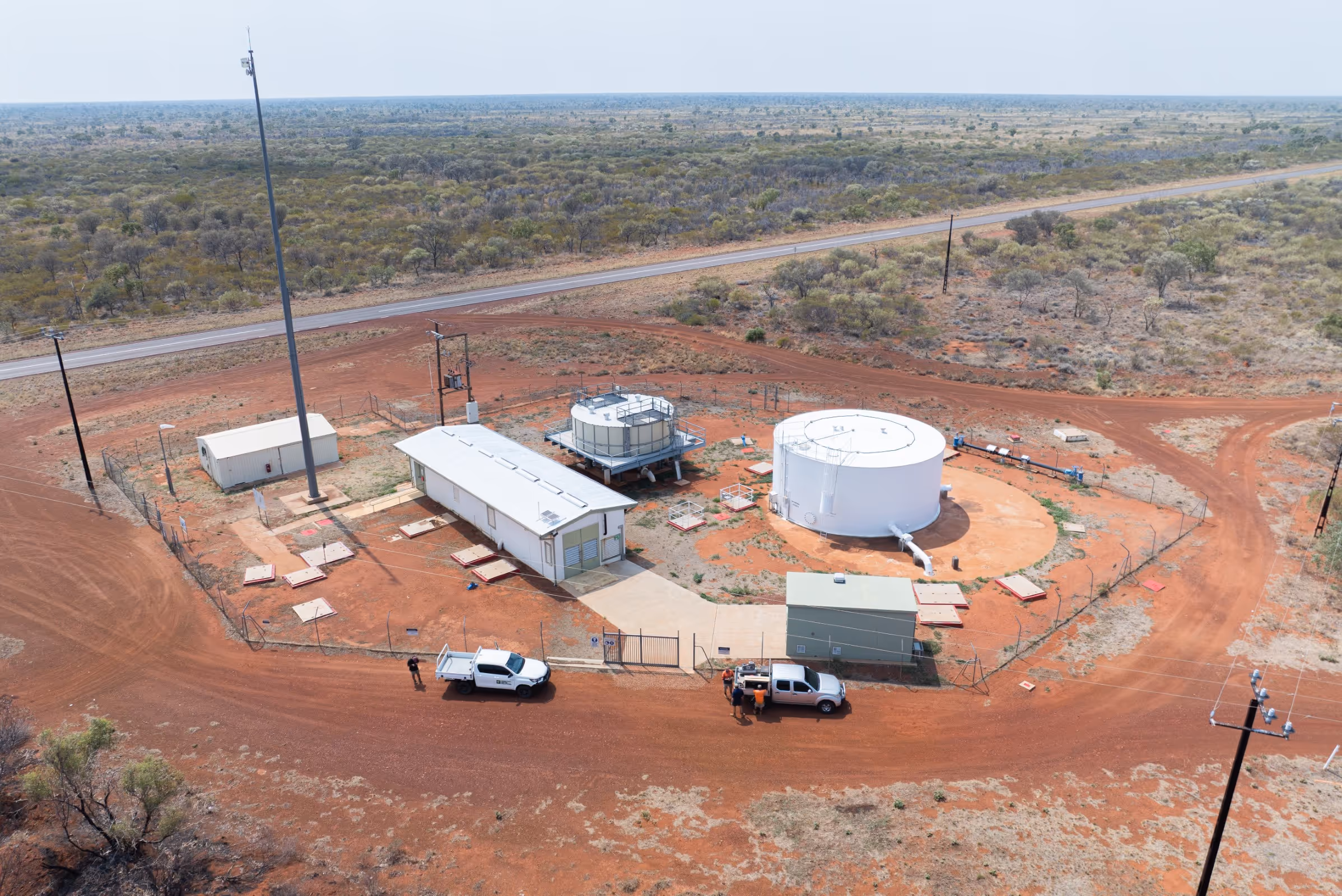 Aerial view of a fenced industrial site with storage tanks, two white utility vehicles, and a small building on red dirt terrain surrounded by sparse vegetation.