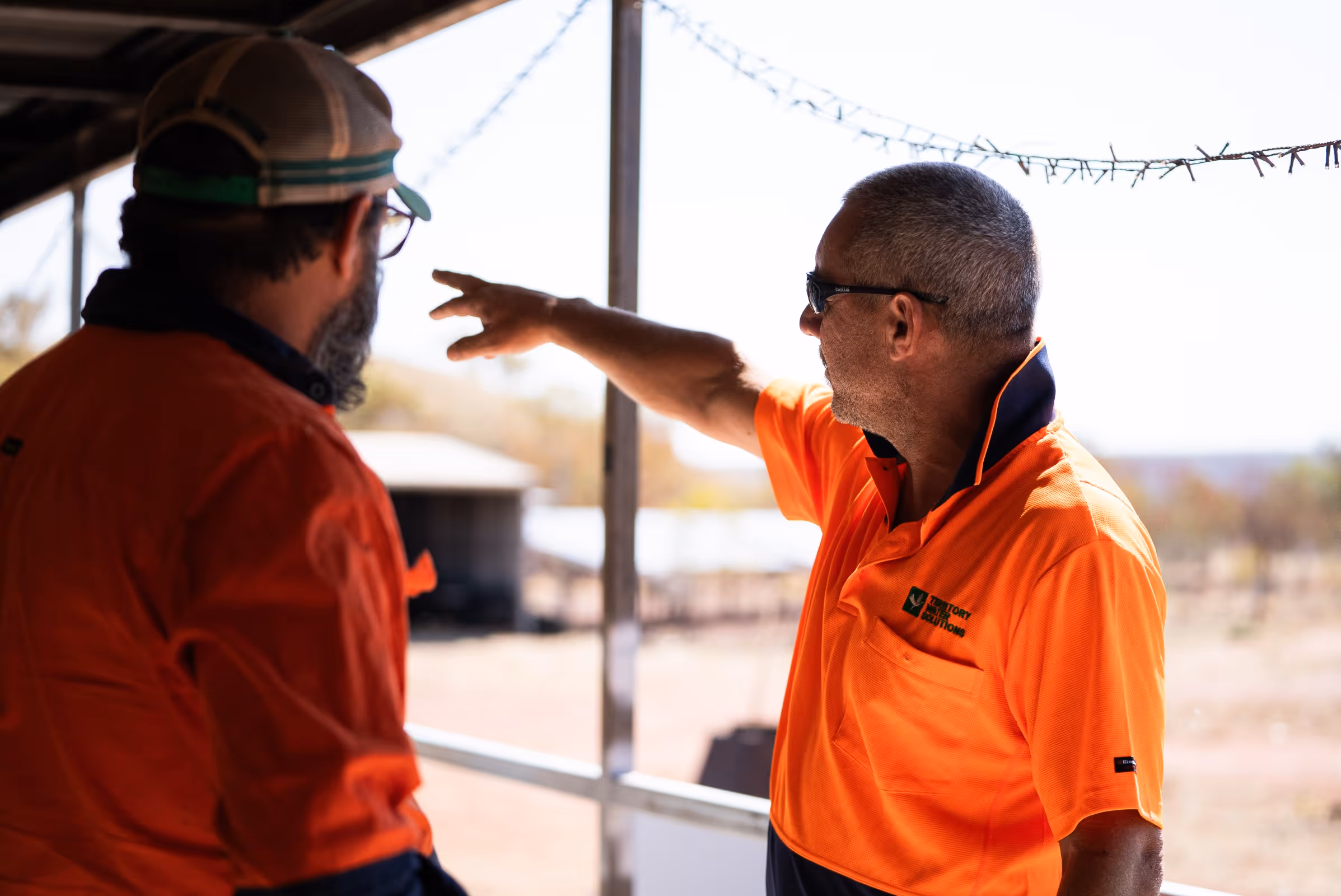 Two men in orange work shirts stand on a porch, with one pointing towards the distance outdoors.