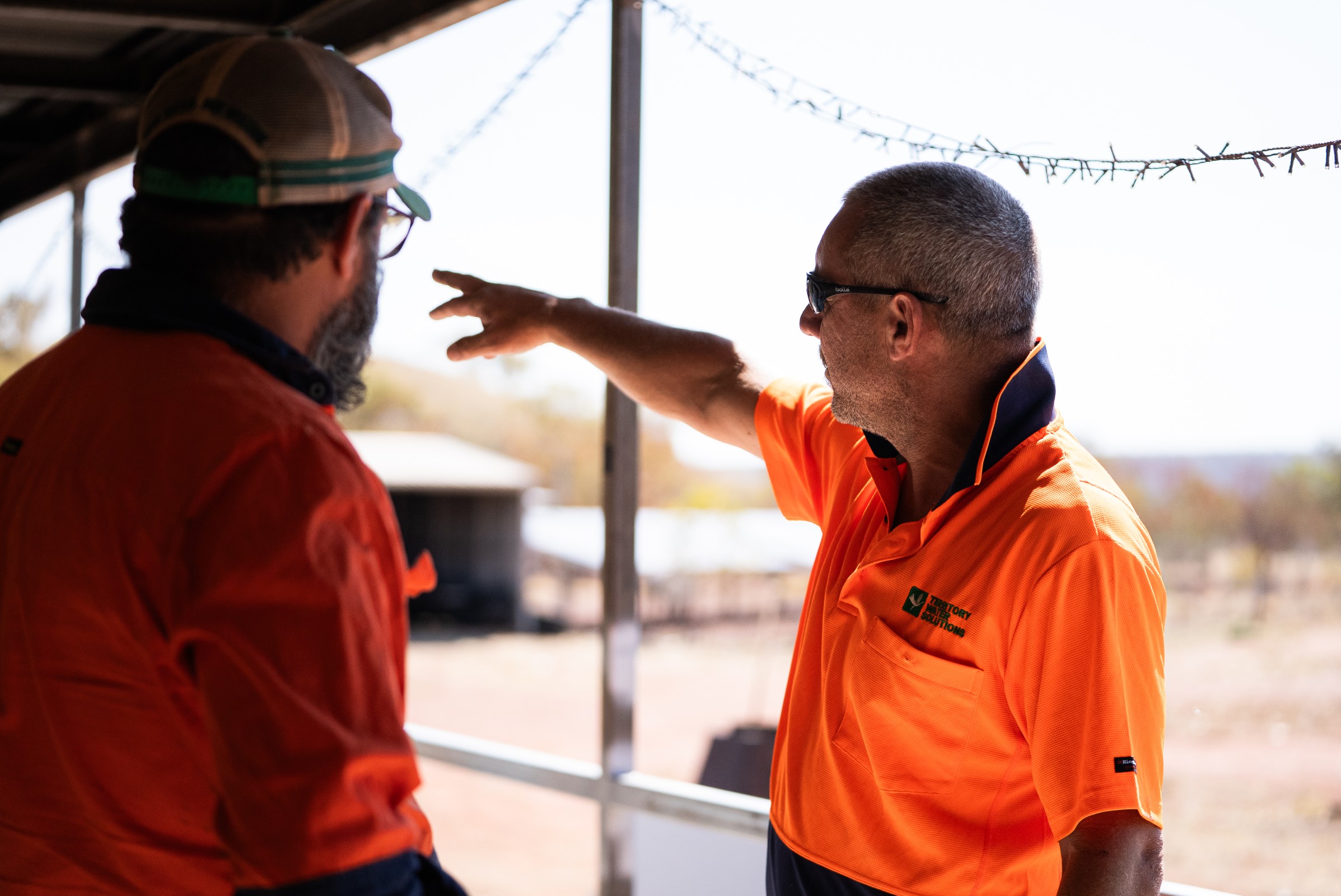 Two men in orange work shirts stand on a porch, with one pointing towards the distance outdoors.
