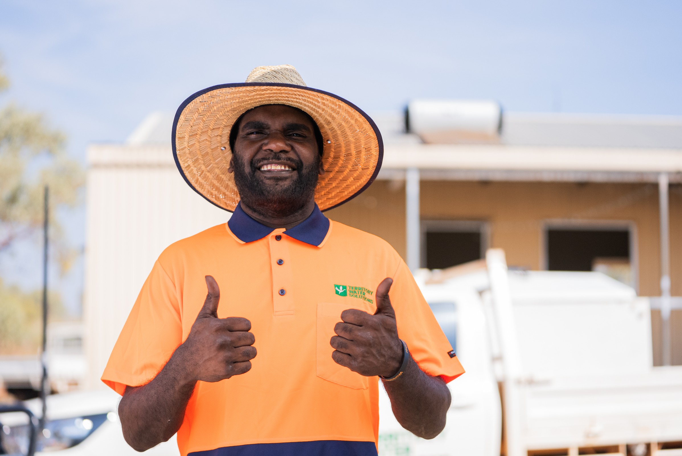 Man wearing an orange safety shirt and straw hat giving two thumbs up outside.