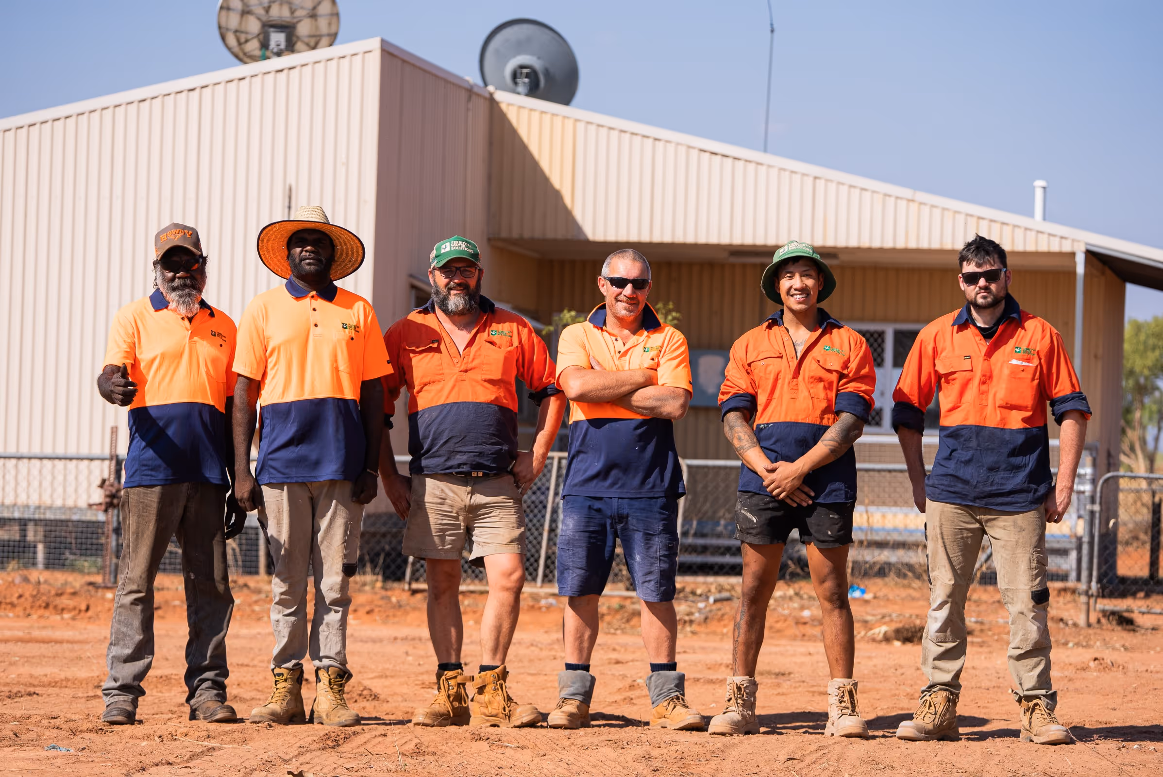 Six construction workers standing side by side outdoors in high-visibility orange and navy uniforms in front of a building.