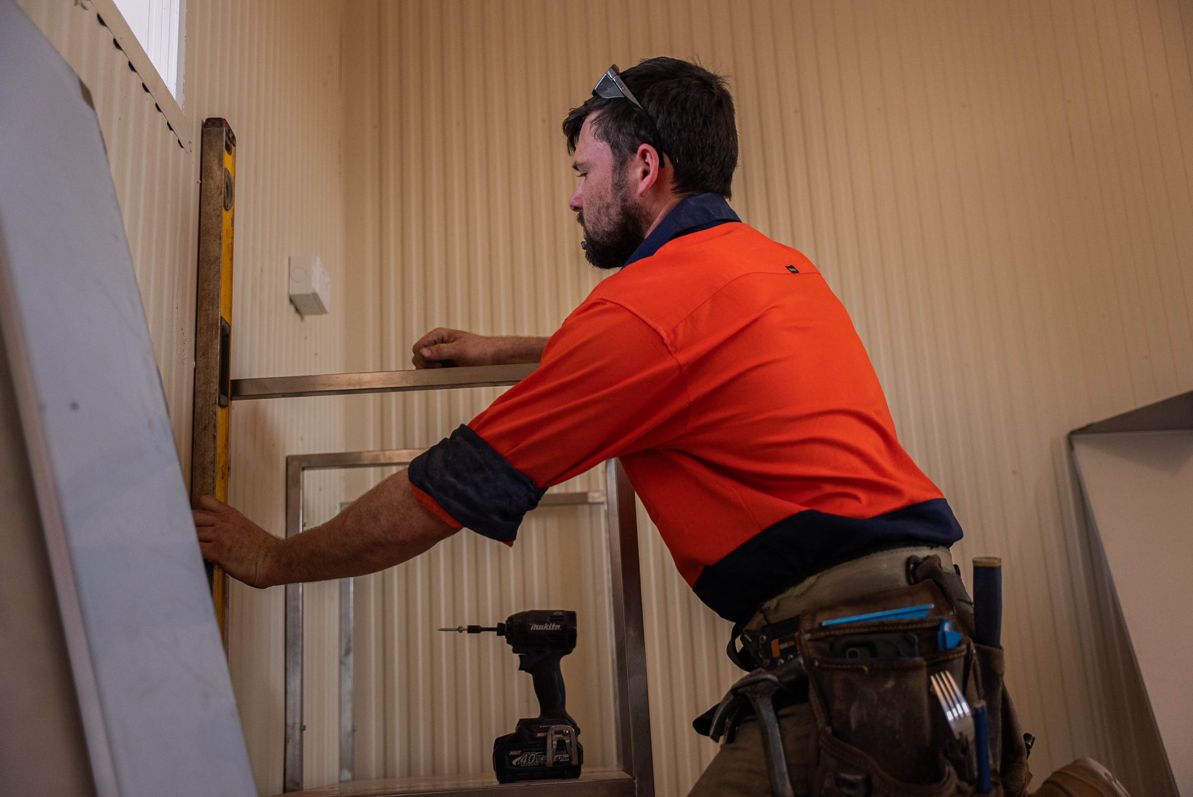 Construction worker in an orange high-visibility shirt using a spirit level on a metal frame indoors.