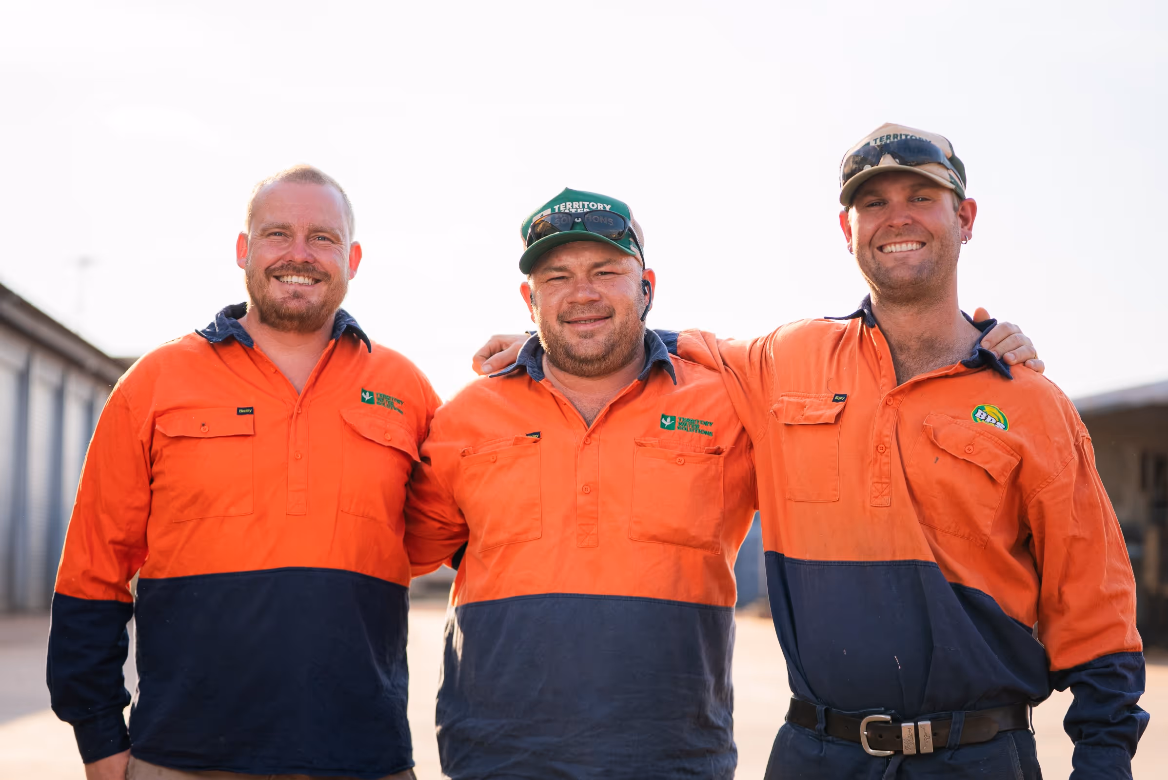 Three smiling men in orange and navy work shirts standing close with arms around each other.