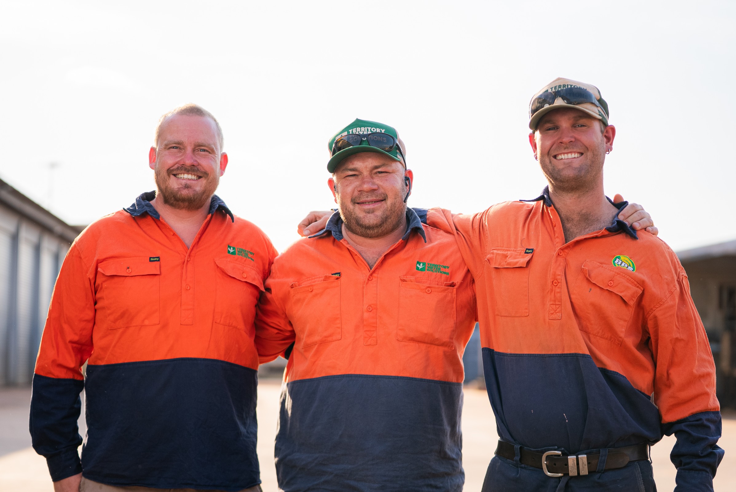 Three smiling men in orange and navy work shirts standing close with arms around each other.