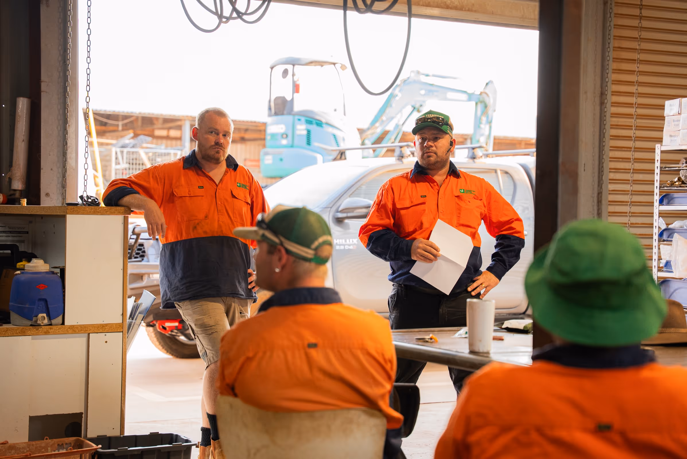 Four construction workers in high-visibility orange and navy shirts having a meeting inside a workshop with machinery and a white pickup truck visible outside.