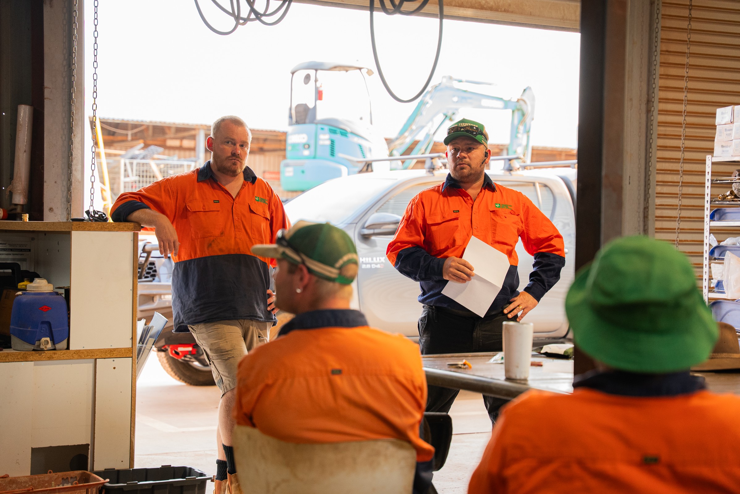 Four construction workers in high-visibility orange and navy shirts having a meeting inside a workshop with machinery and a white pickup truck visible outside.