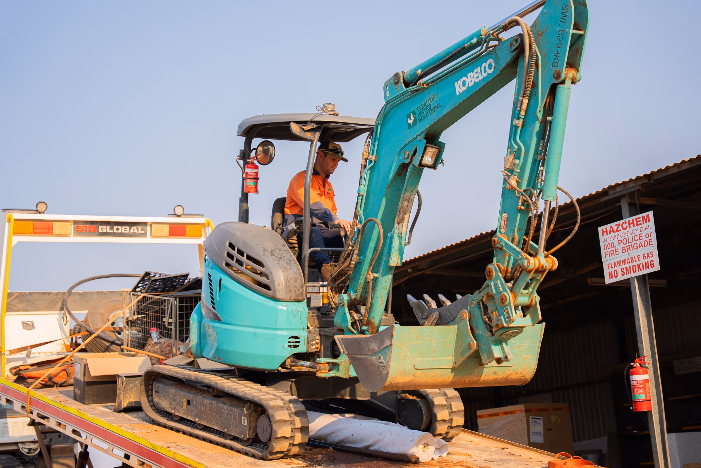 Construction worker operating a teal Kobelco mini excavator on a flatbed trailer next to a hazard sign and fire extinguisher.