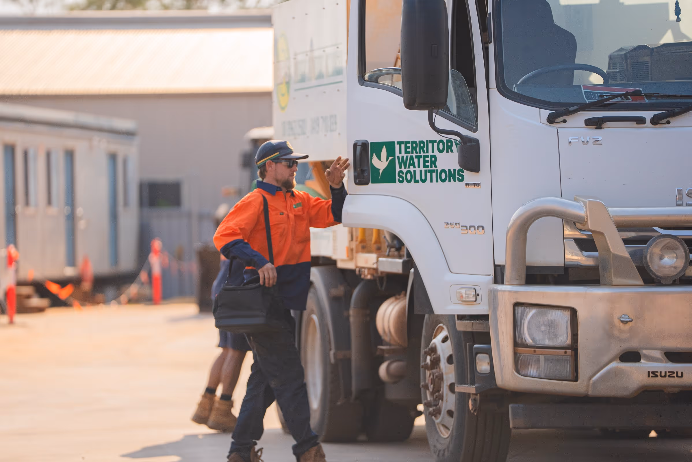 Worker in orange and navy uniform carrying a black bag walking next to a white Territory Water Solutions truck.