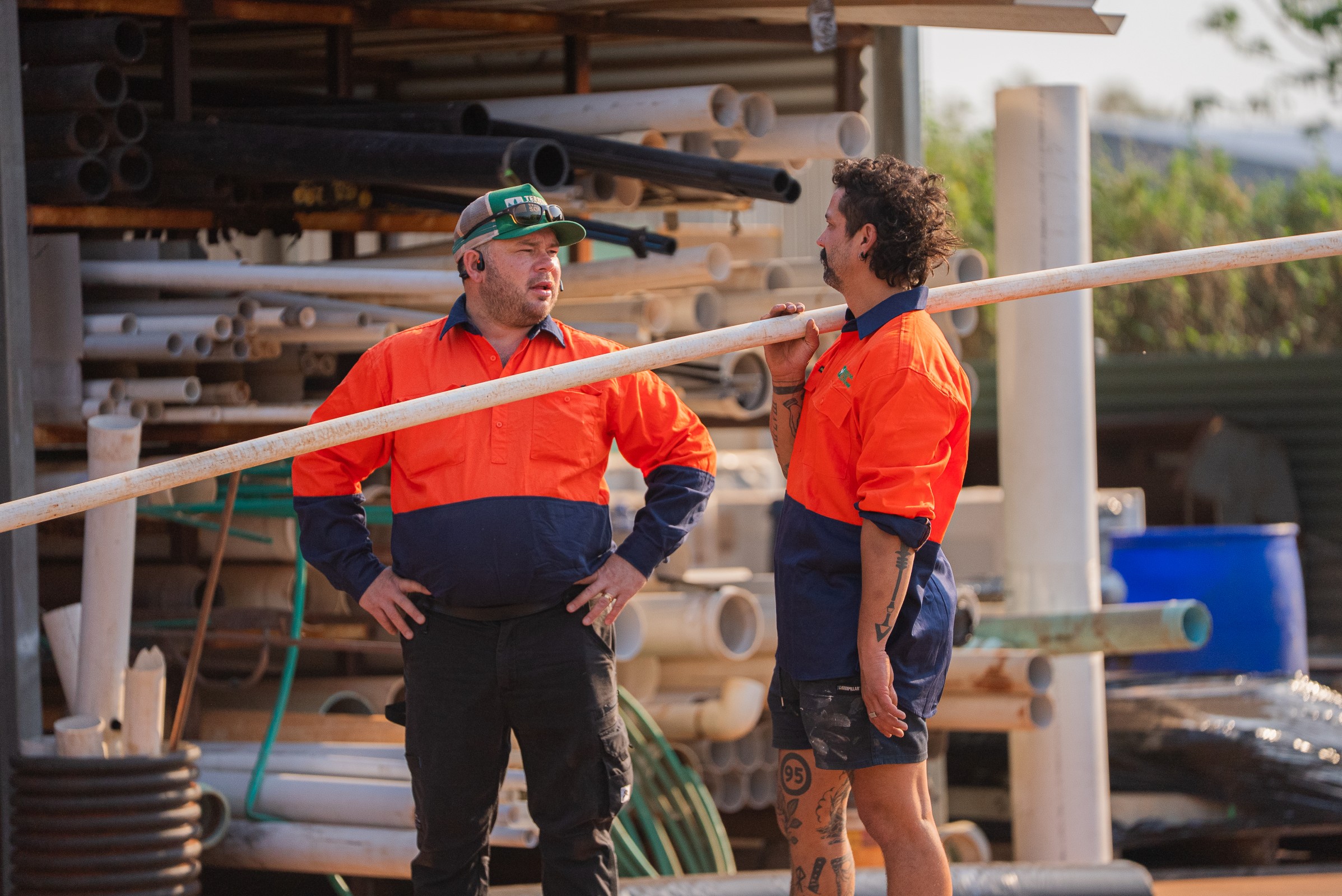 Two construction workers in orange and navy uniforms holding a long pipe and talking at a pipe storage yard.