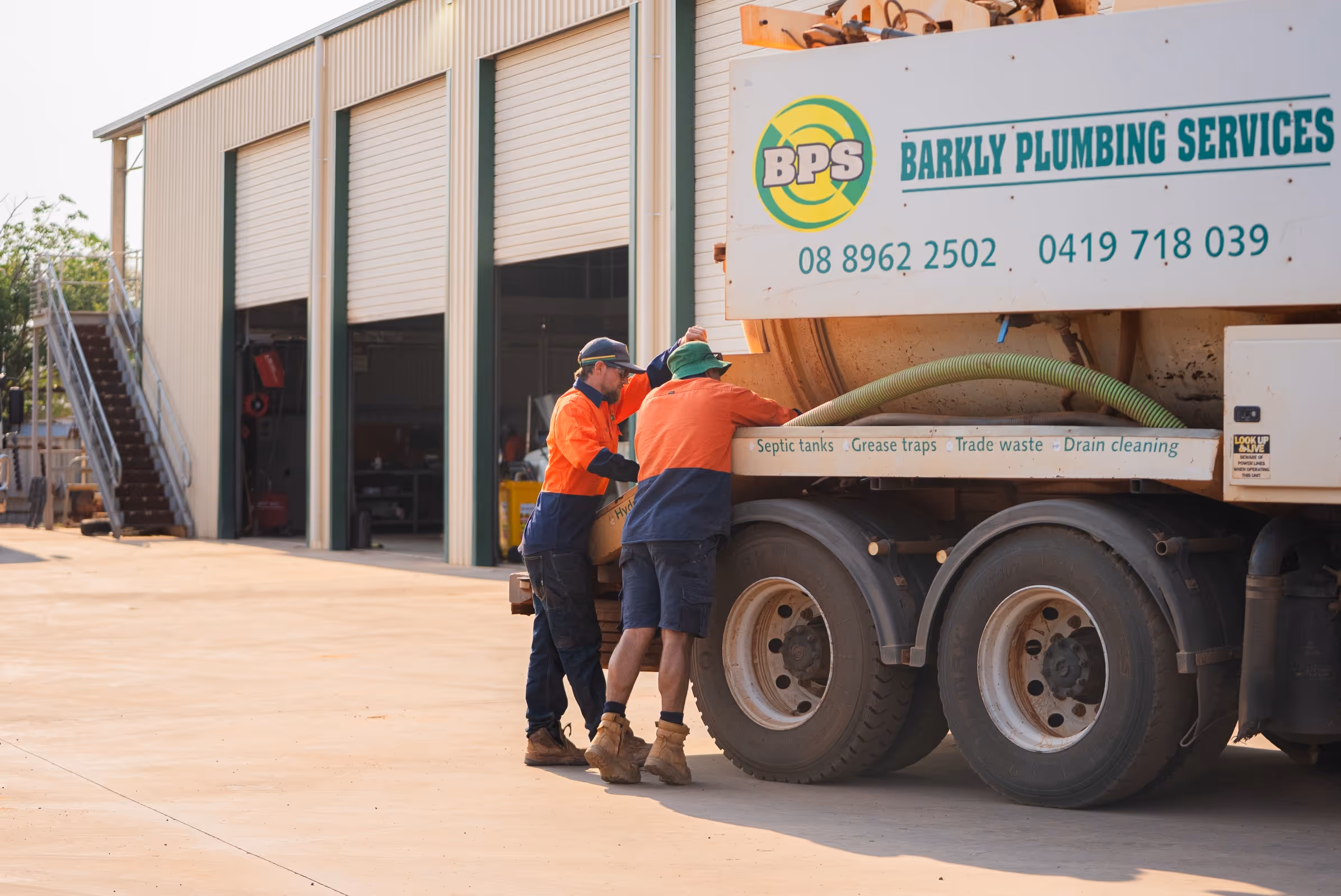 Two workers in orange and navy uniforms inspecting a Barkly Plumbing Services septic tank truck outside a warehouse.