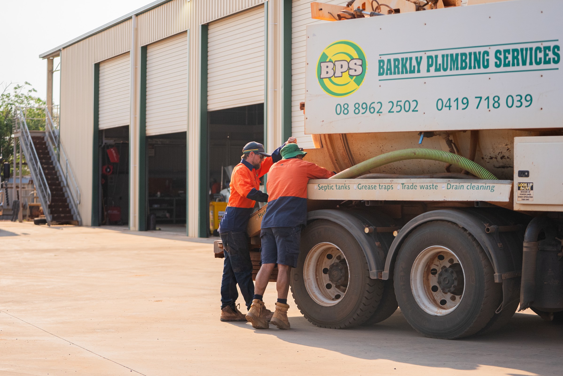 Two workers in orange and navy uniforms inspecting a Barkly Plumbing Services septic tank truck outside a warehouse.