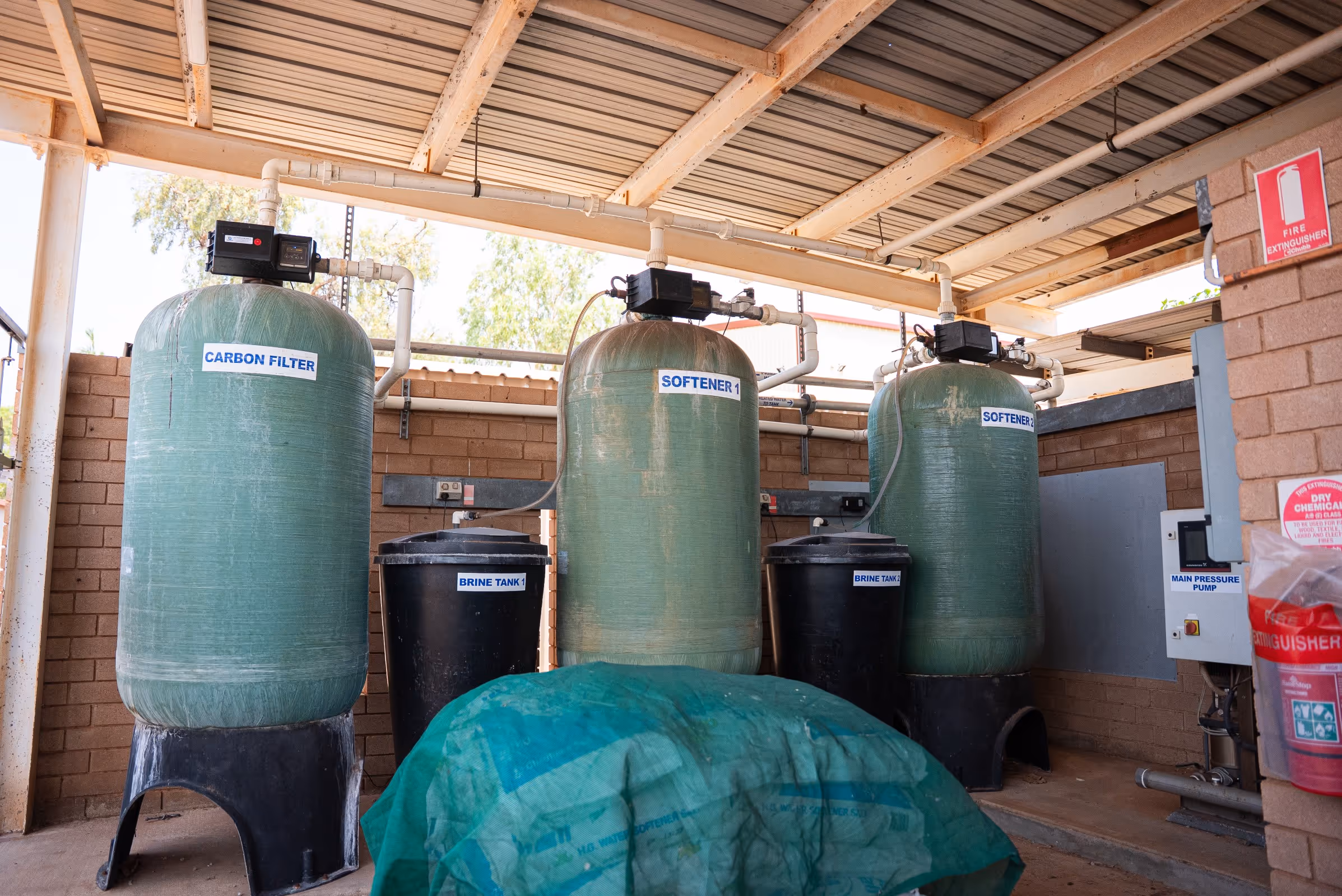 Three large green water treatment tanks labeled Carbon Filter, Softener 1, and Softener 2, each paired with black brine tanks, in an industrial setting with a fire extinguisher visible on the right wall.